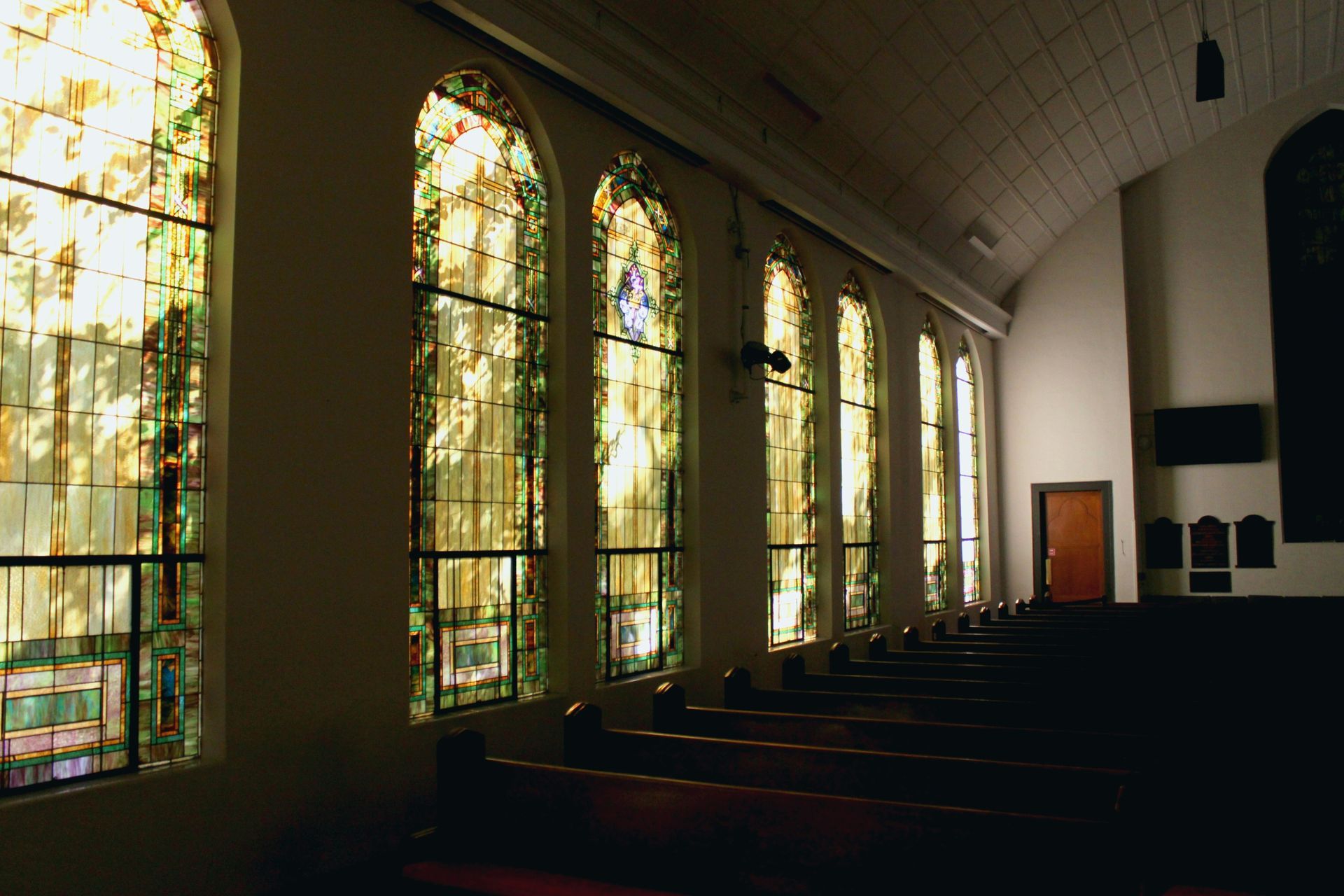 Interior church view with stained glass windows along one side, wooden pews, and a door at the end.