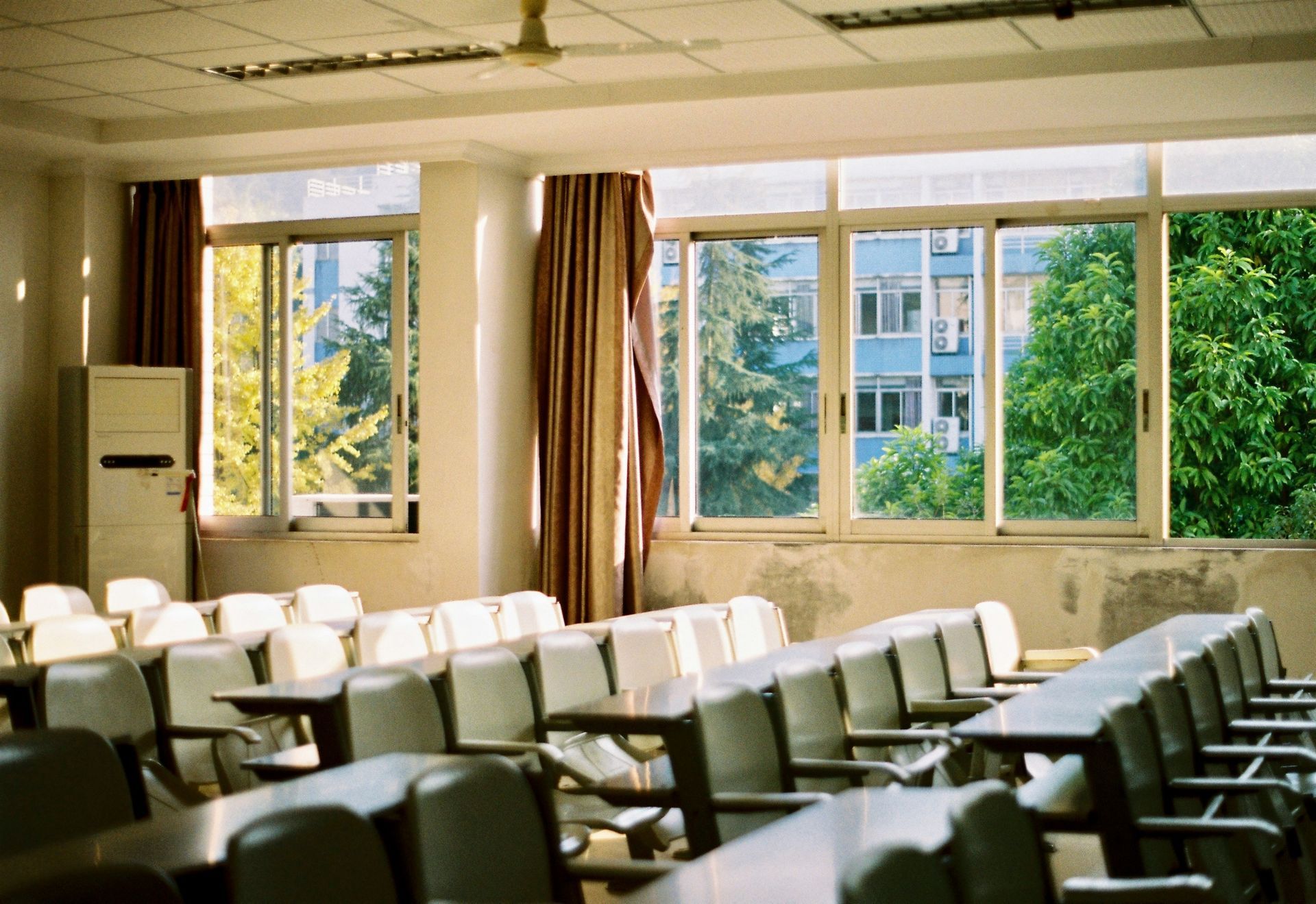 Empty classroom with rows of desks facing windows overlooking trees and a building.