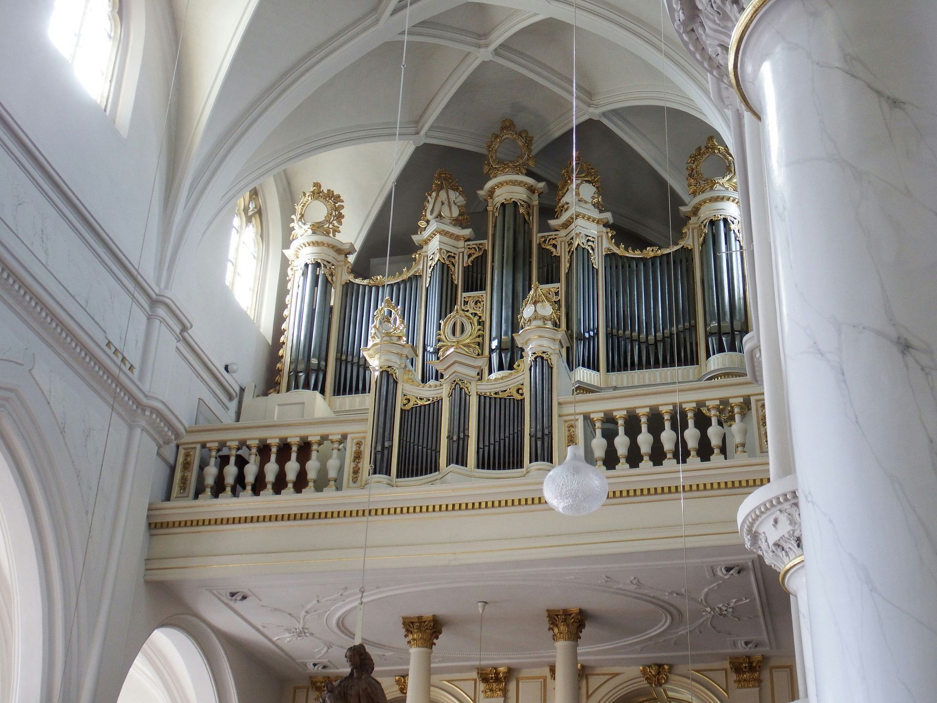 Ornate pipe organ on a balcony inside a light-filled church. White and gold accents, tall columns.