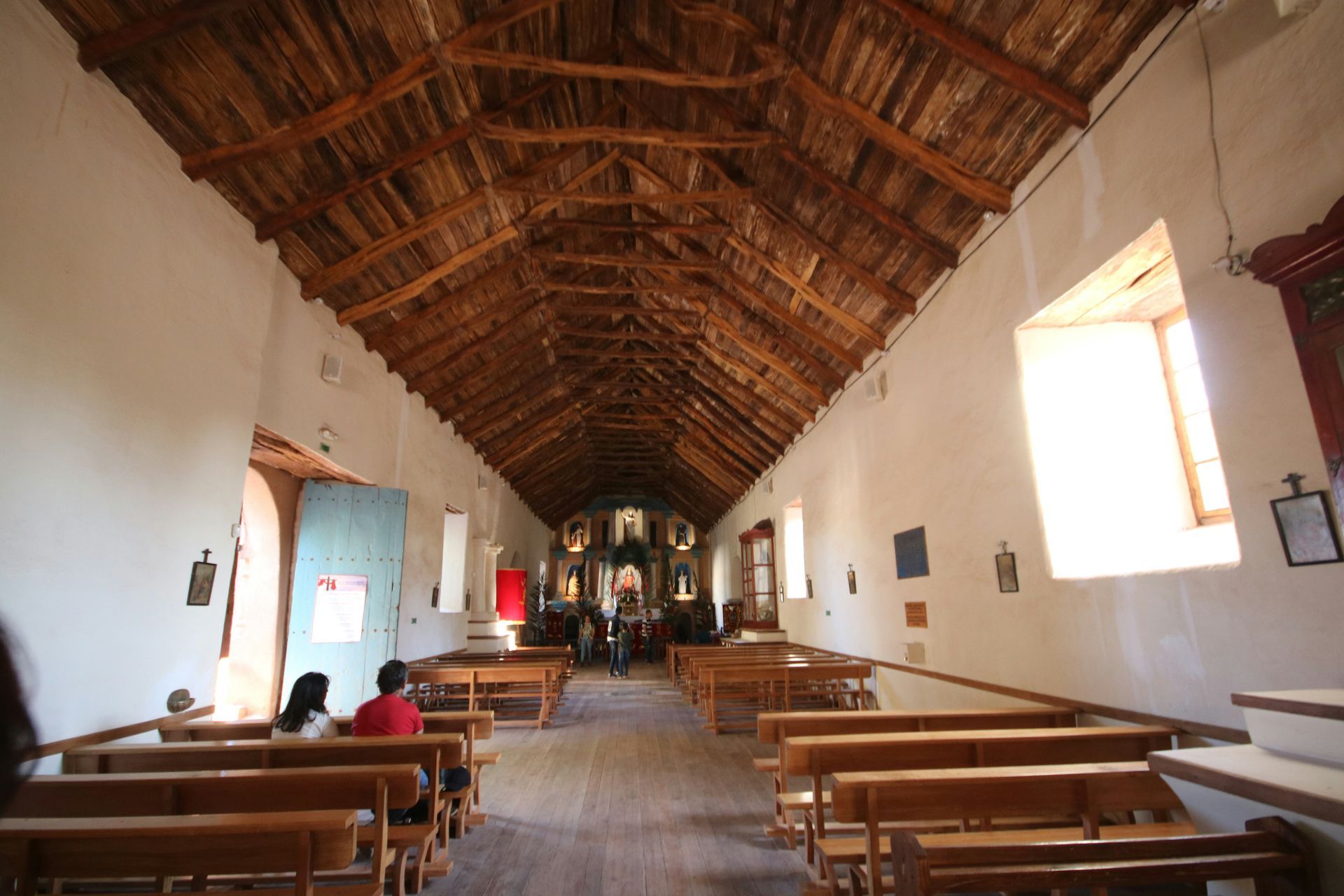 Interior of a historic church with wooden beams, white walls, and rows of pews. Two people sit in the foreground.