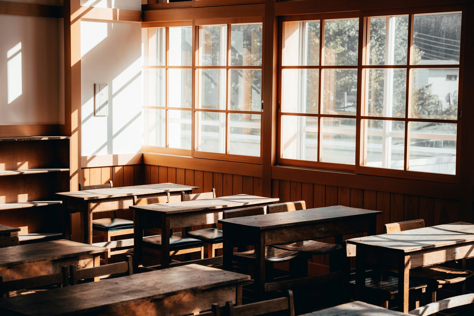 Empty sunlit classroom with rows of wooden desks and large windows.