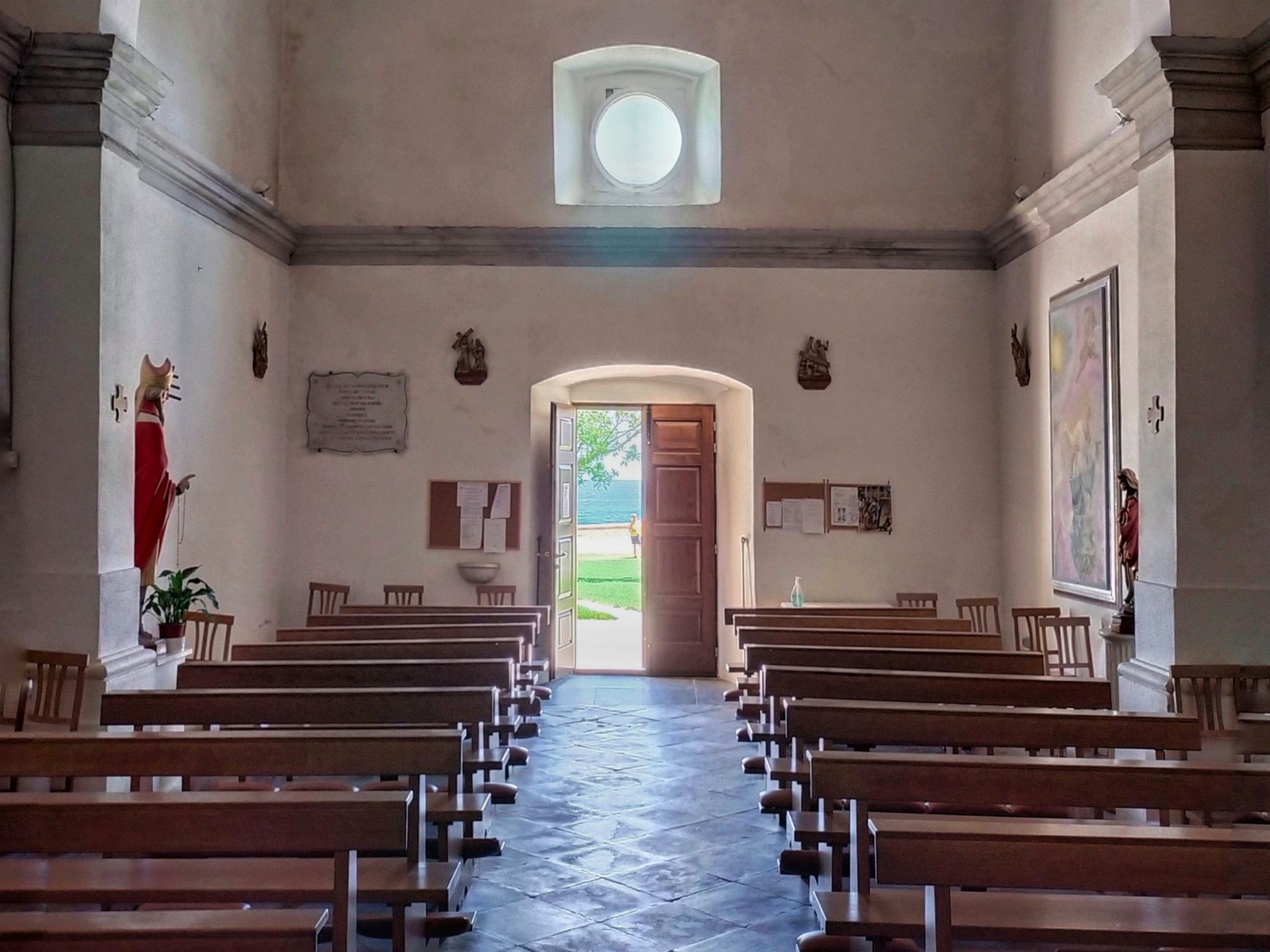 Interior of a church with rows of wooden pews leading to an open doorway with sunlight streaming in.