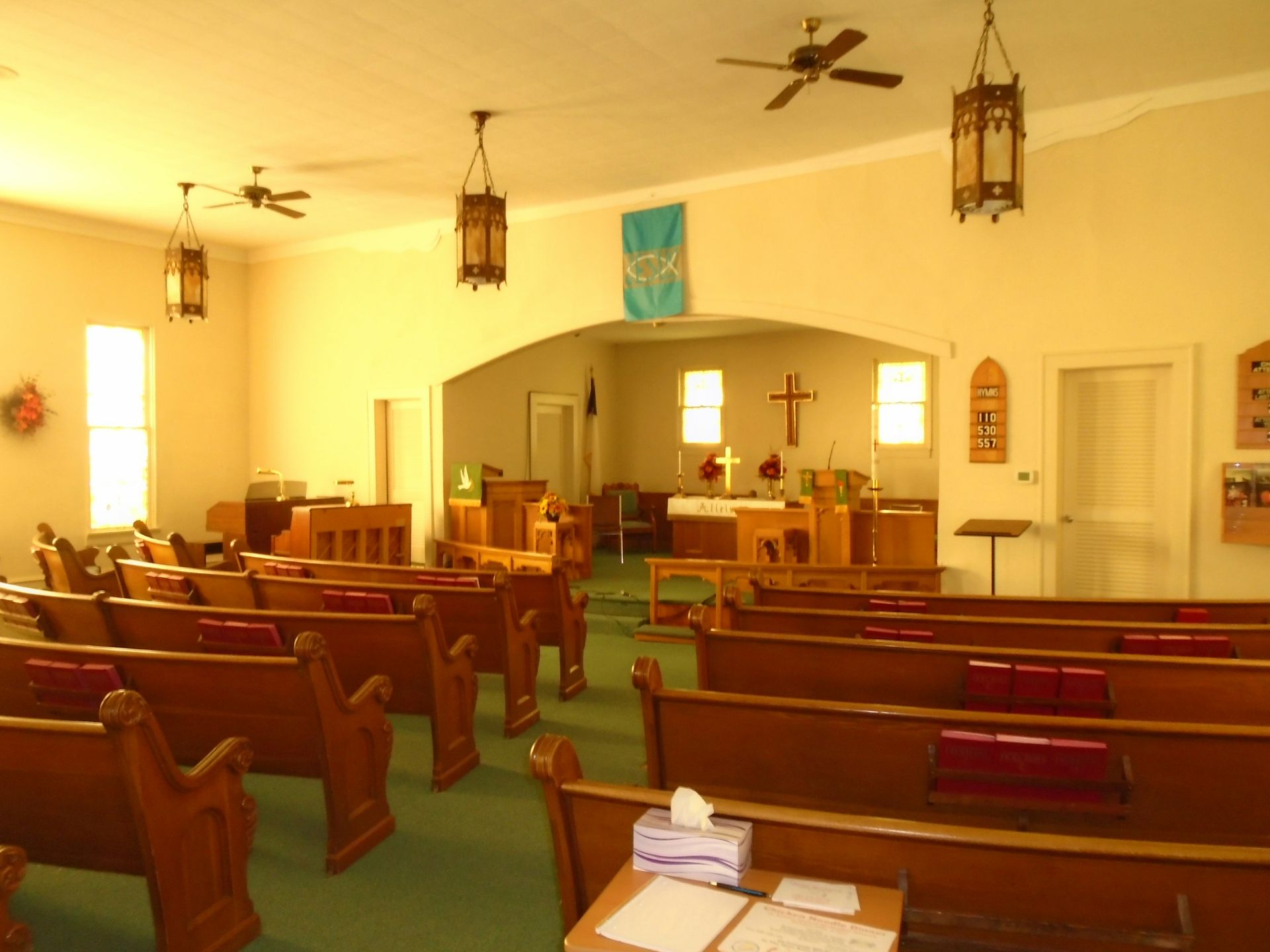 Interior of a church sanctuary with wooden pews, altar, and hanging lamps; archway leads to chancel area.