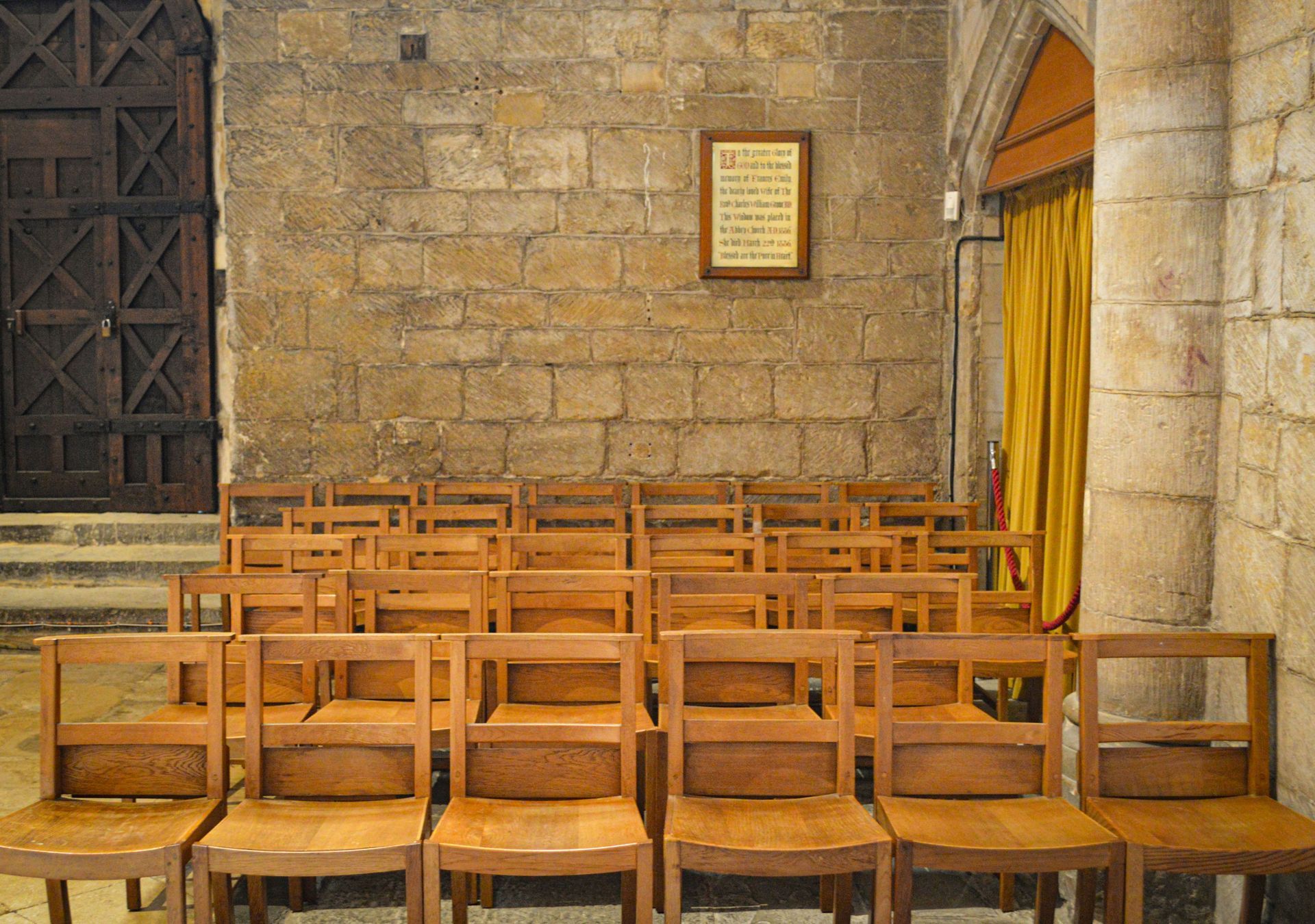 Rows of wooden chairs face the interior of a stone building. A framed document hangs on the stone wall.
