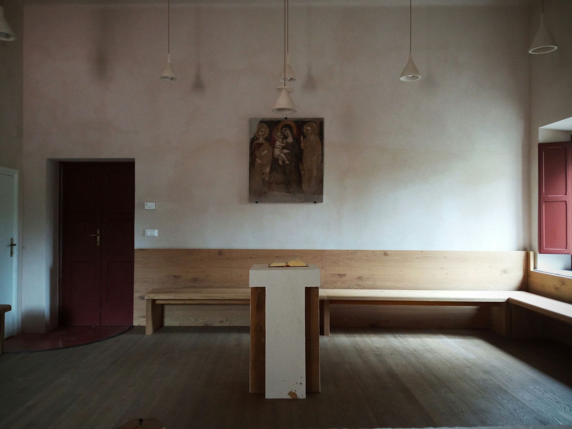 A simple, wooden interior with benches, a podium, and a painting on the wall. Red door and window shutters.