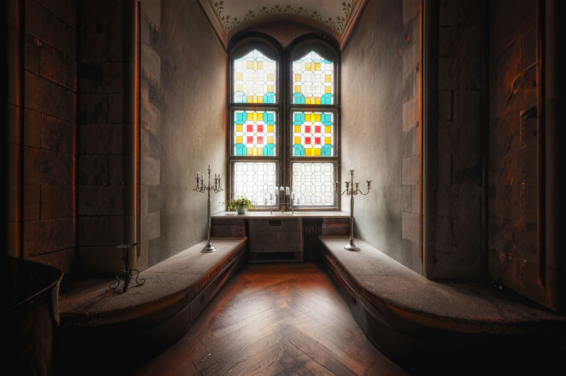 Window alcove with stained glass, stone benches, and candelabras, in a dimly lit space.