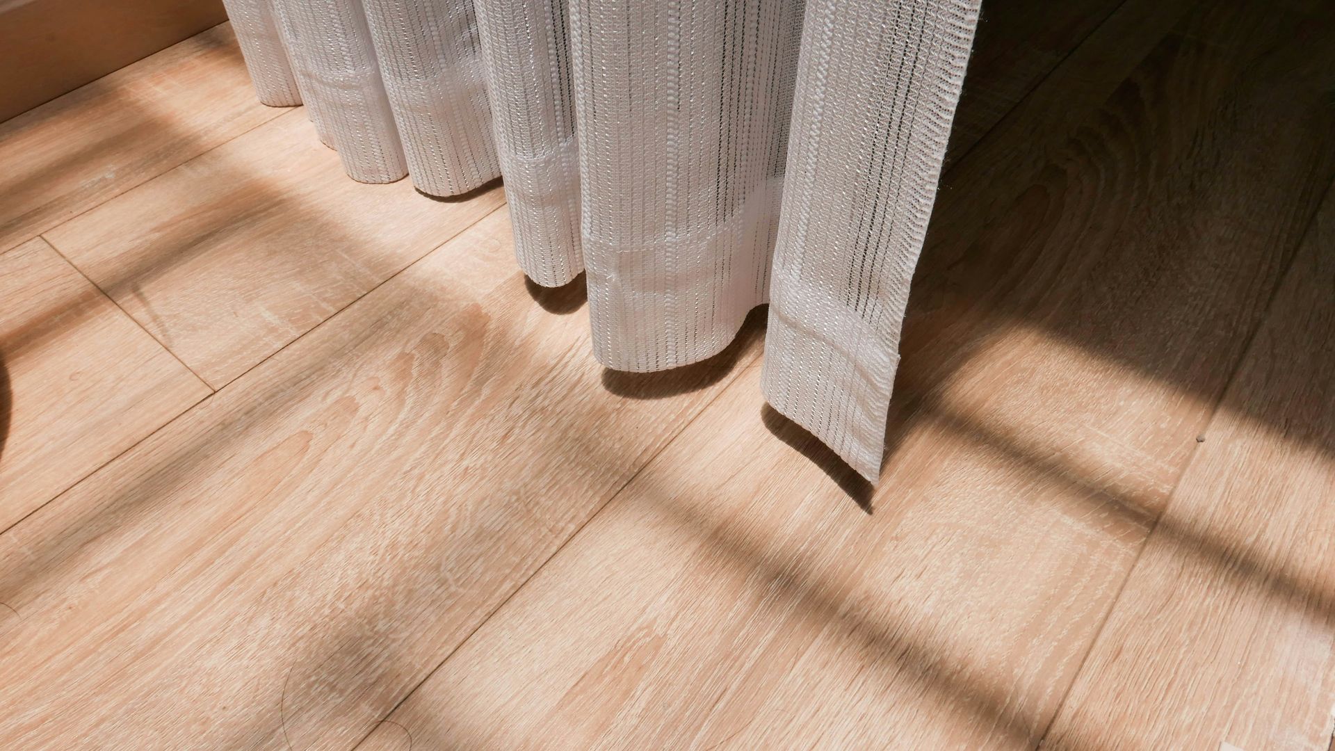Curtain's hem casting shadows on light wood-look floor. Sunlight streams through, creating stripes.