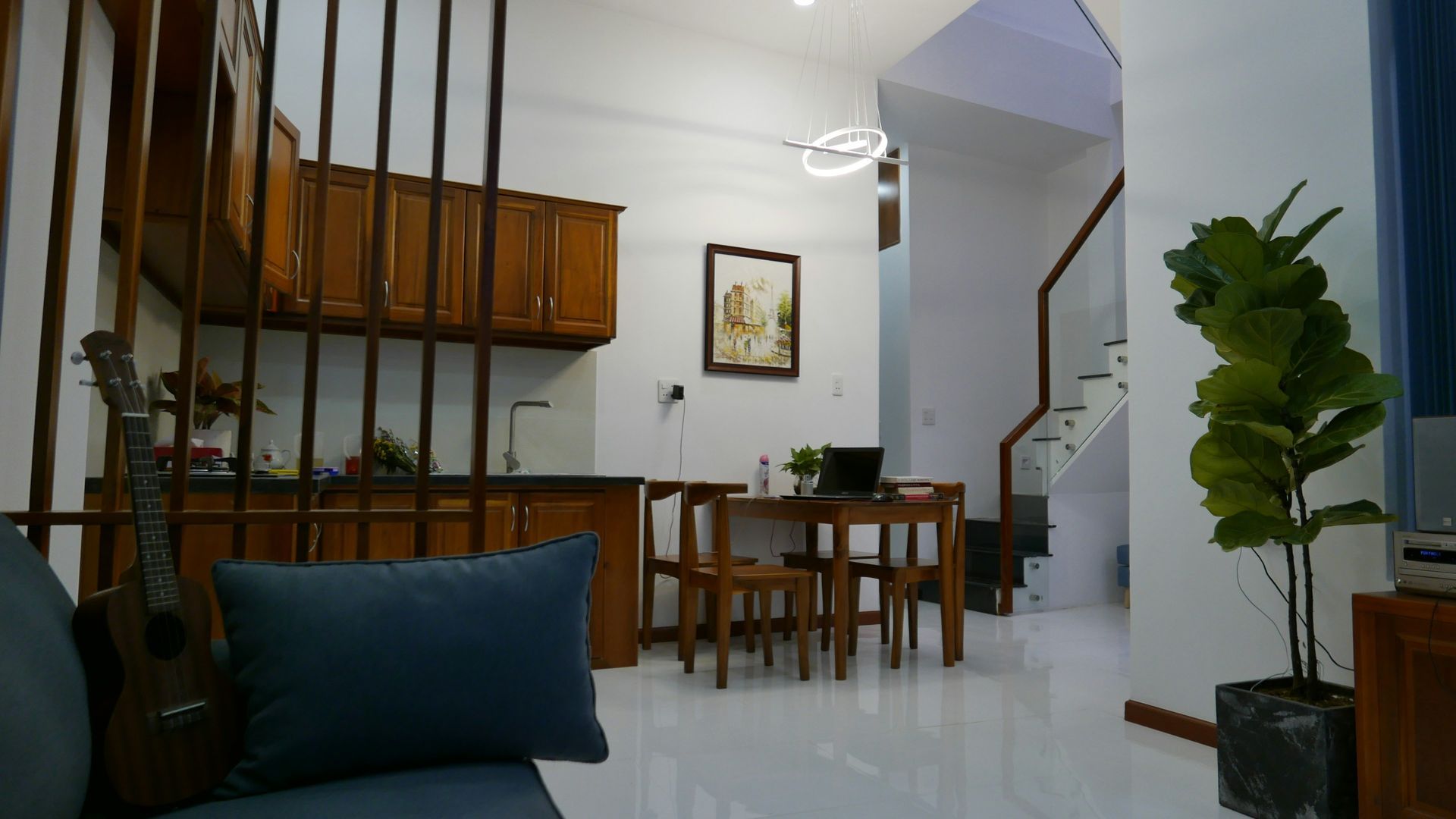Interior view of a kitchen and dining area with wooden cabinets, table, and a staircase. A large potted plant sits nearby.