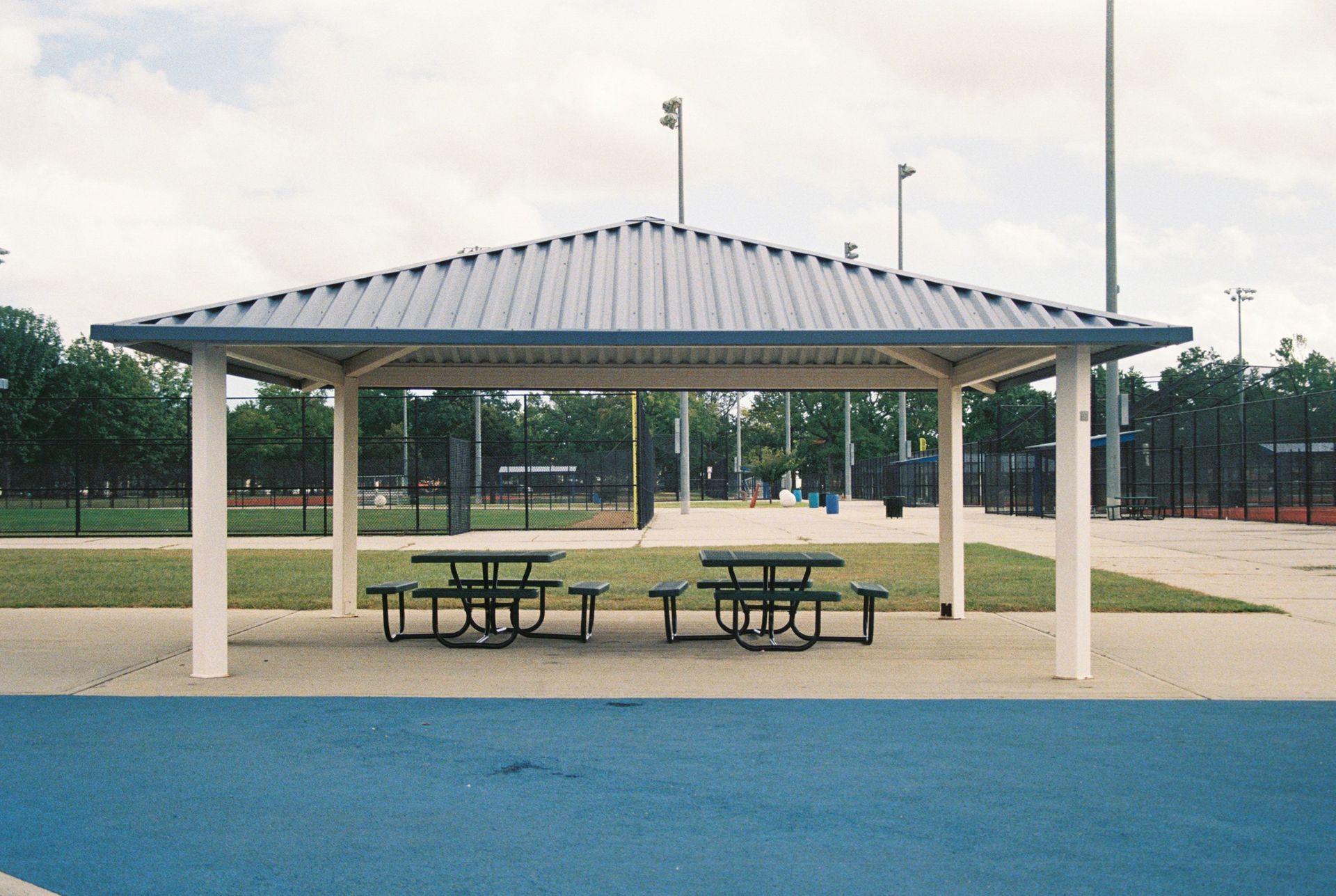 Picnic shelter with two tables on a blue surface, near a grassy area and sports fields.