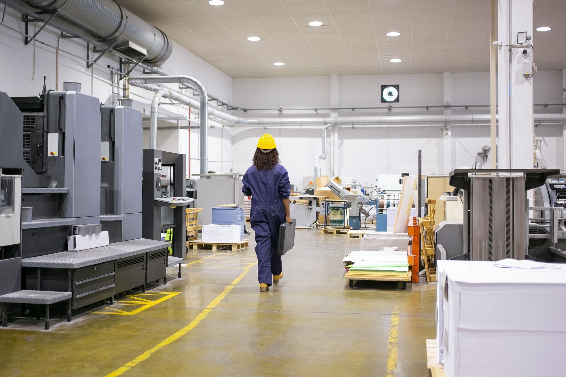 Person in a blue jumpsuit and yellow hard hat walks through a factory holding a briefcase.