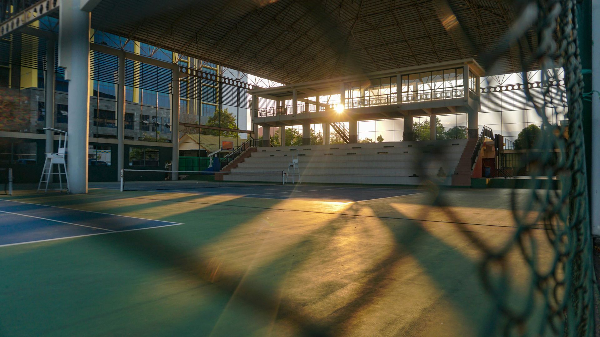 Indoor sports court with sunlight streaming through a roof structure; blurred fence in foreground.