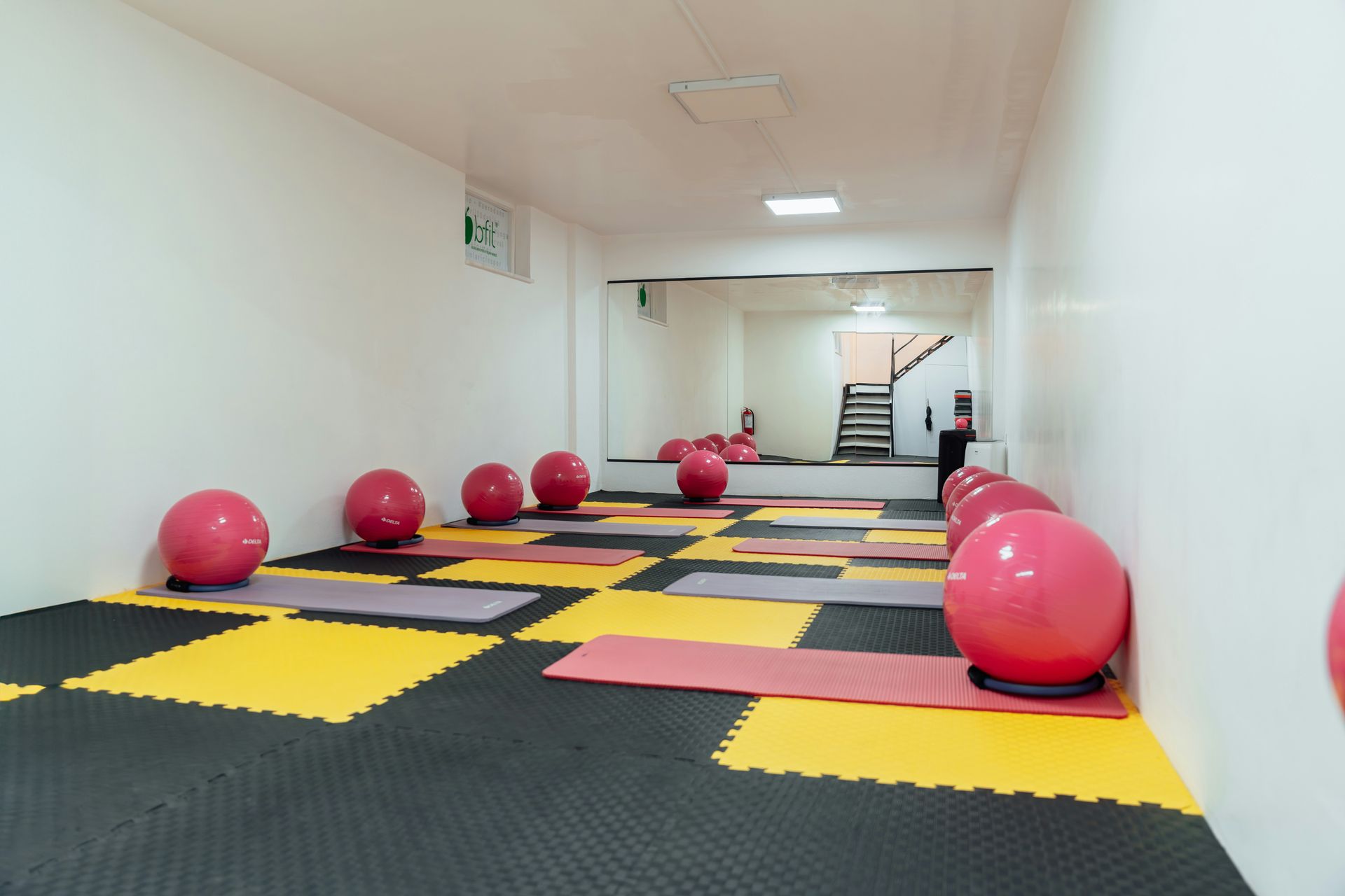 Gym space with pink exercise balls, mats on a black/yellow floor, mirrored wall.