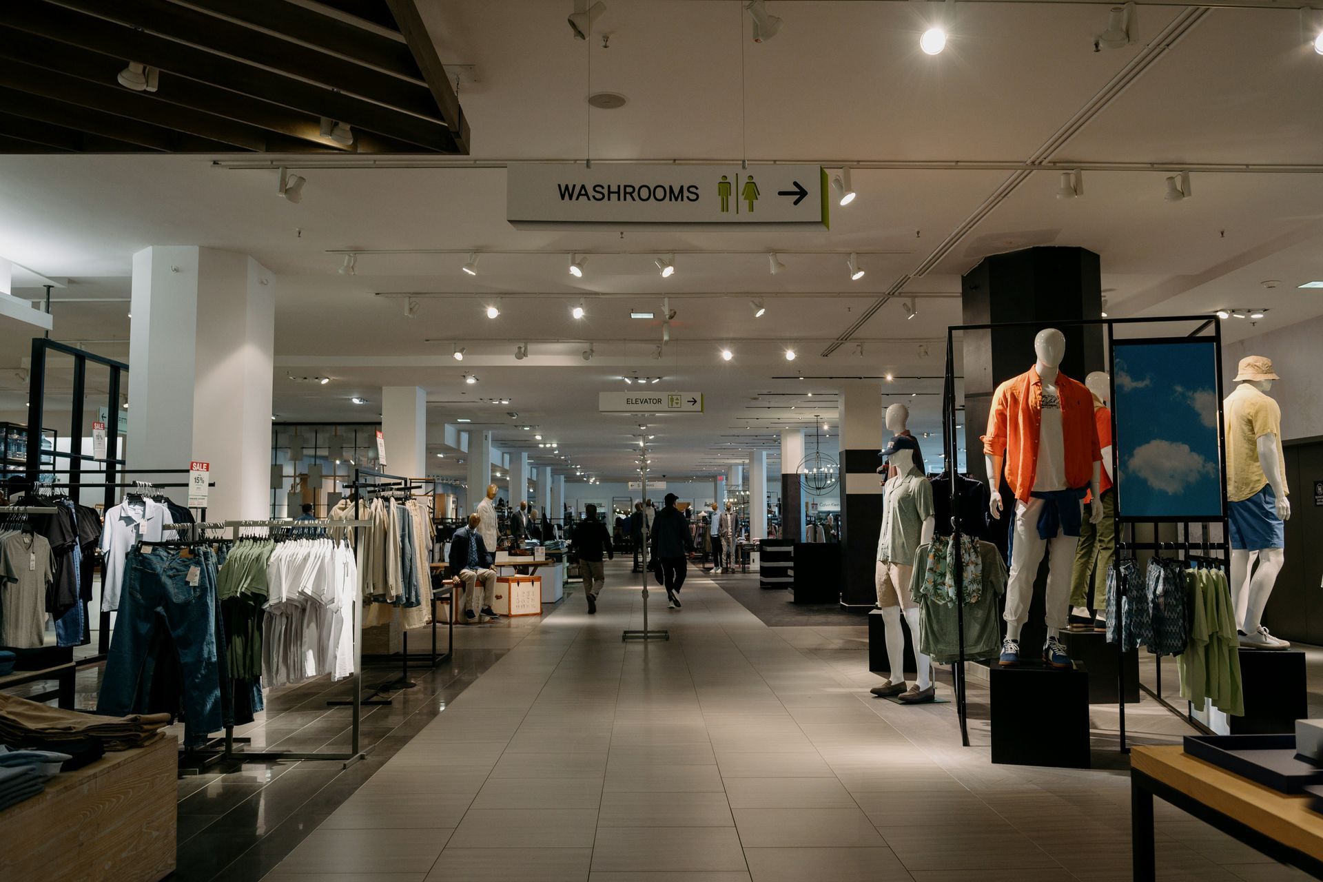 Inside a clothing store, mannequins and racks of clothes line the long hallway. Overhead directional sign.