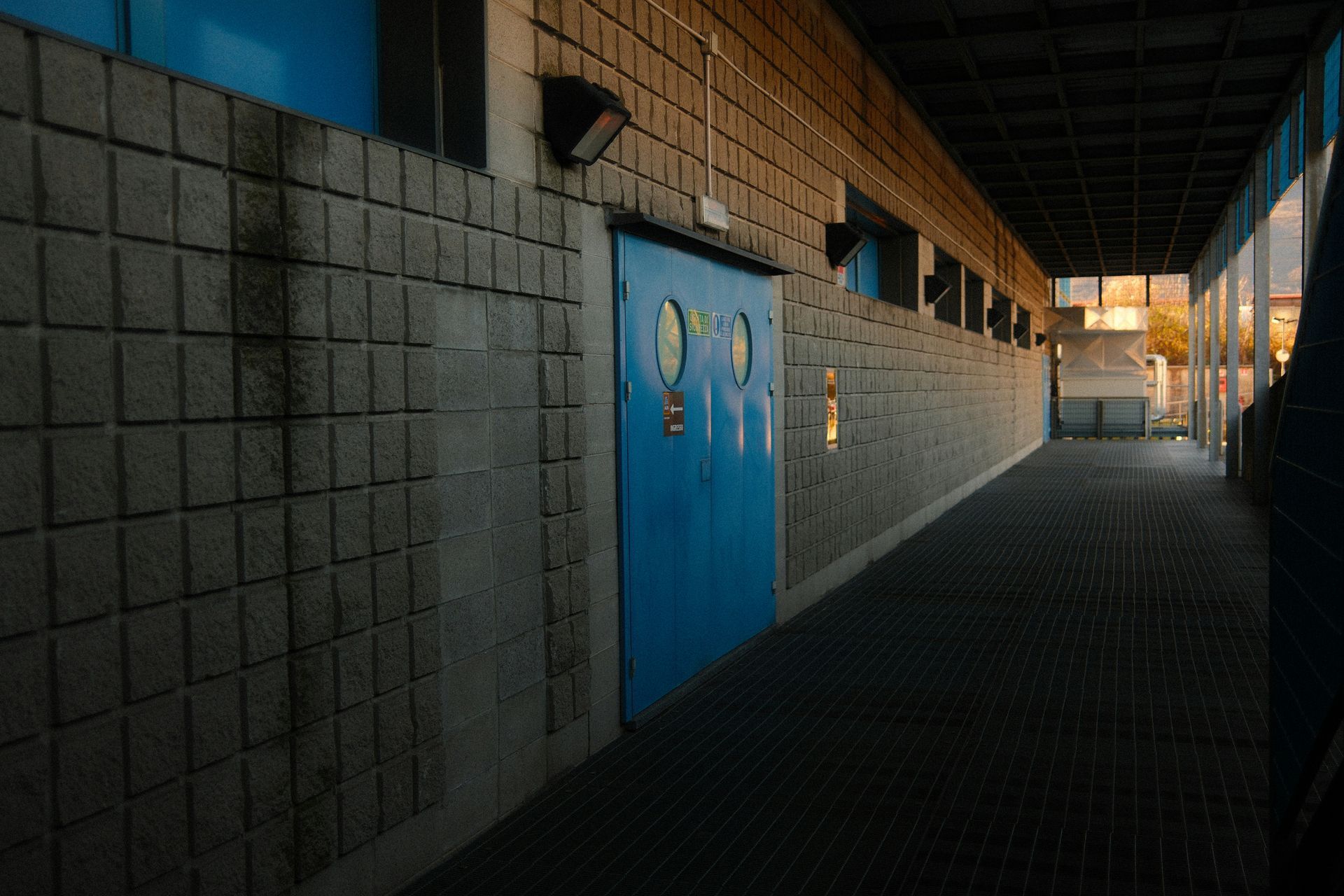 A dim, brick-walled hallway with a blue door and circular windows, bathed in sunlight at the end.
