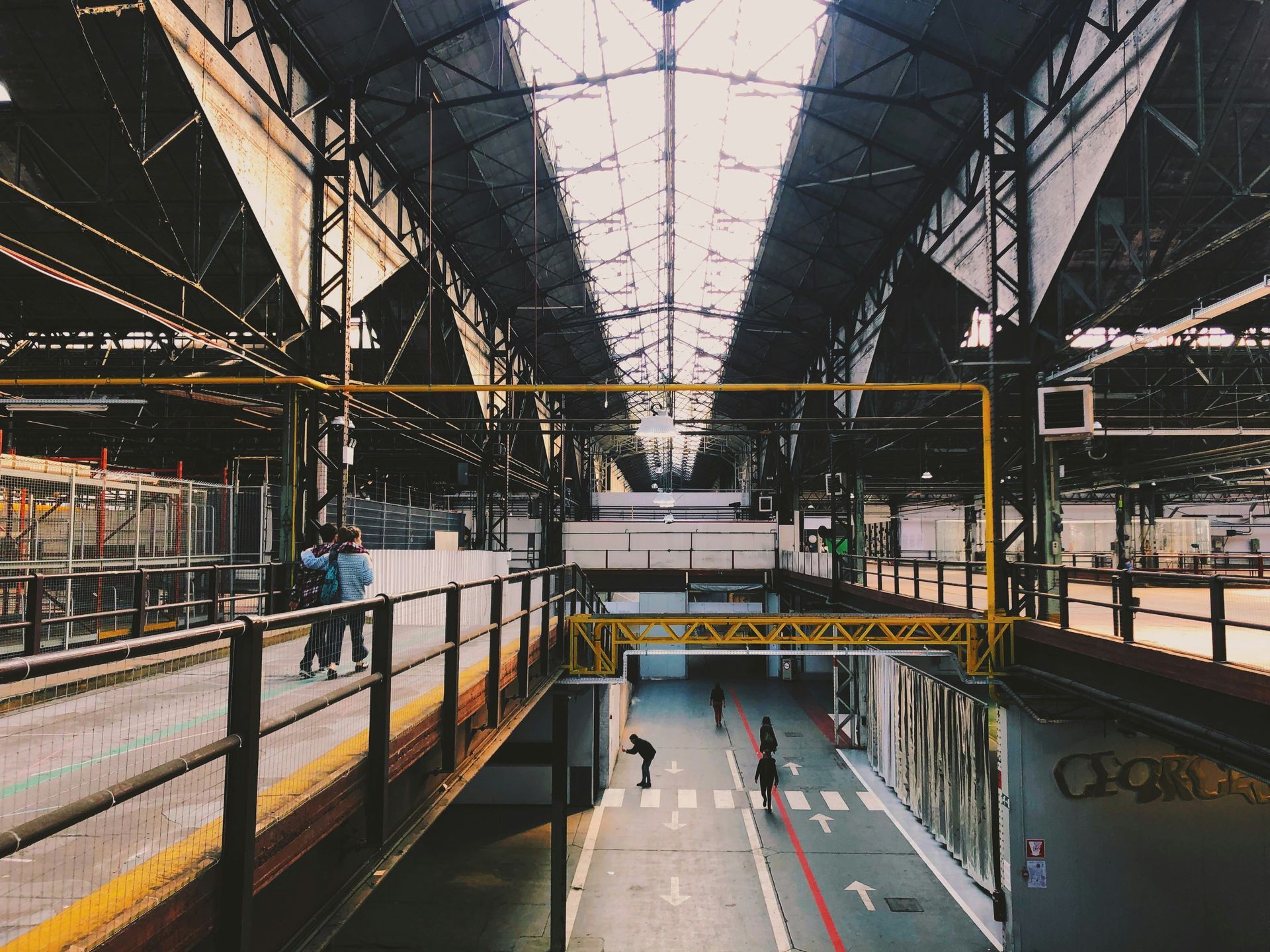 Interior of an industrial building. People walking on a walkway above a large open space with a metal roof.