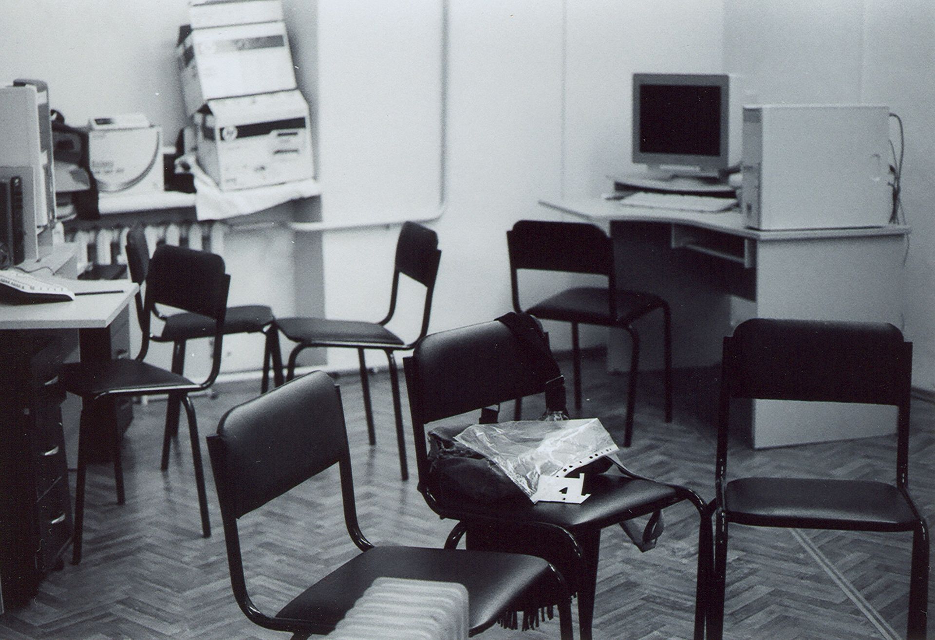 A messy room with chairs, a desk with a computer, and boxes stacked against a wall.