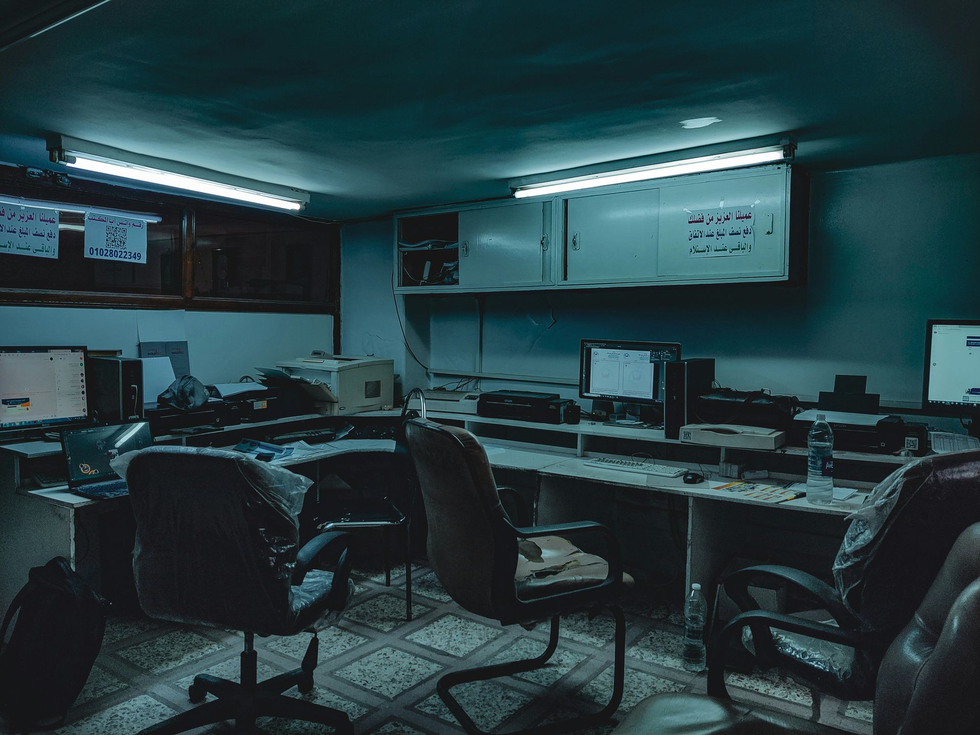 Dimly lit, cluttered computer lab with desks, chairs, monitors, and a whiteboard.