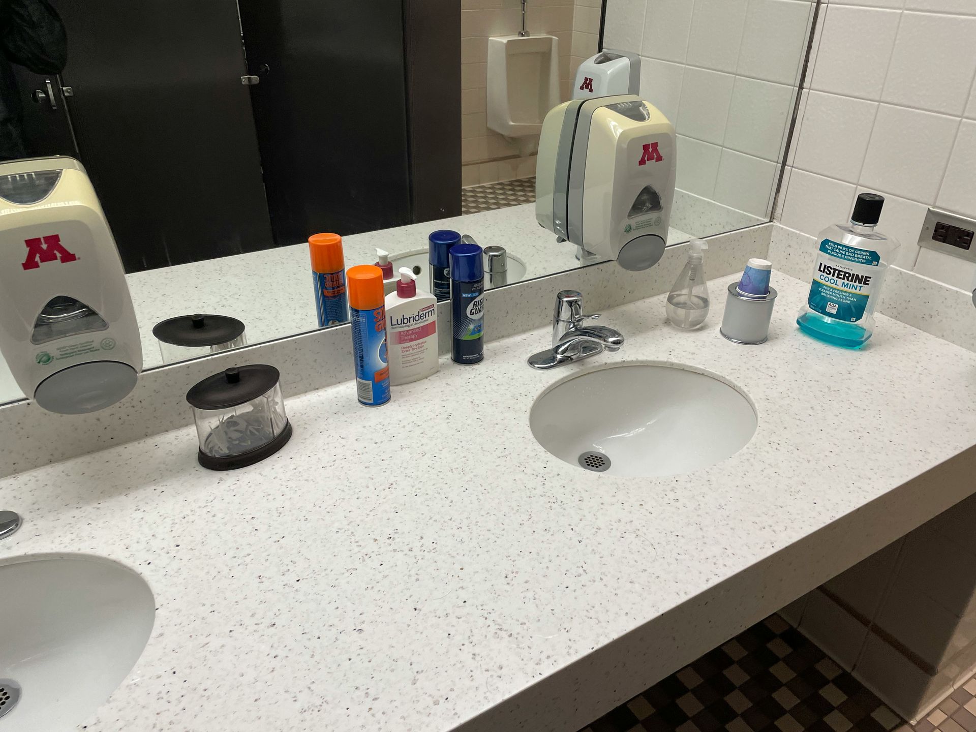 Public restroom sink counter with soap dispensers, mirror, and various toiletries.