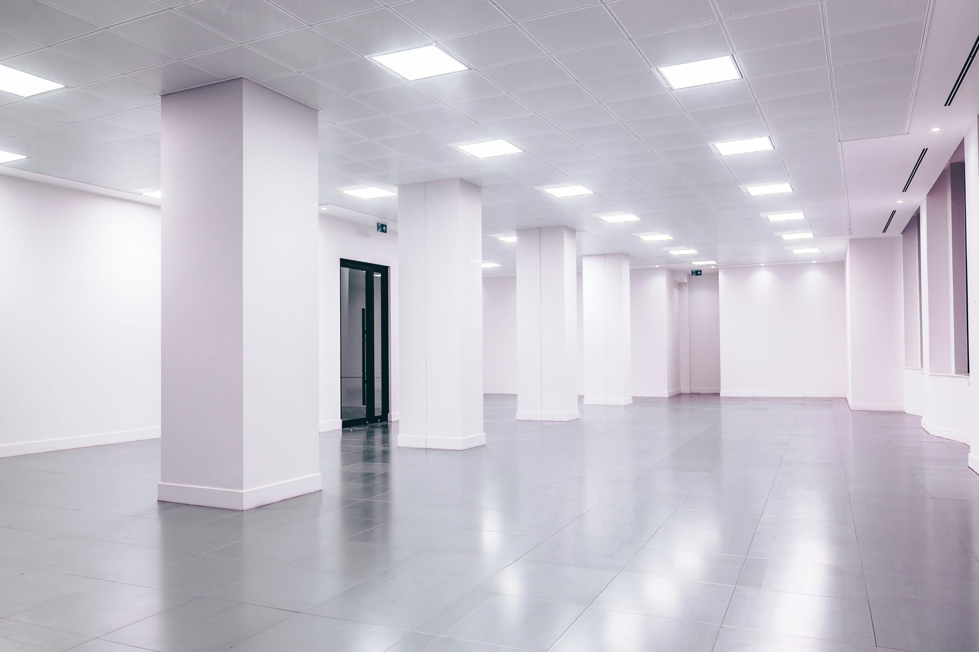 Empty, bright, modern room with columns and a polished floor. Fluorescent lights and a doorway are visible.