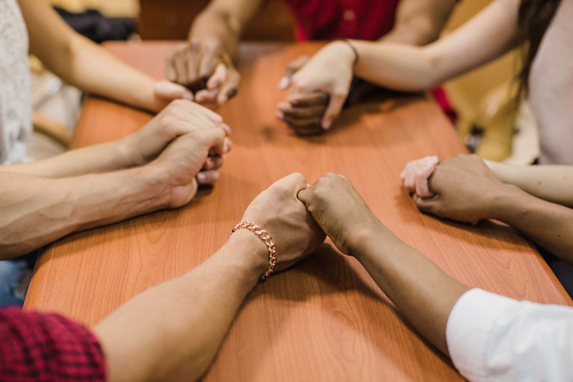 Hands clasped in prayer around a wooden table. Diverse group.