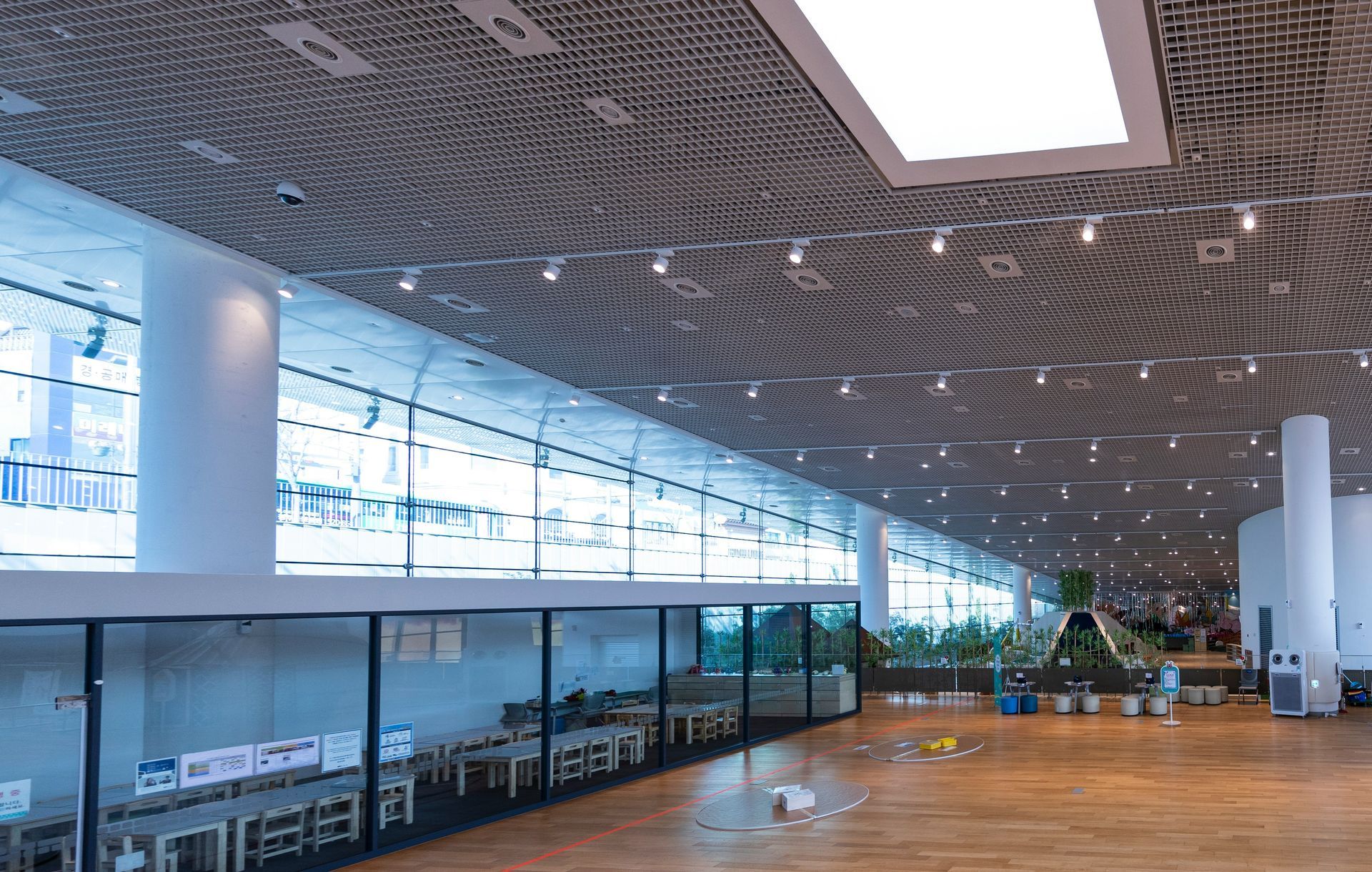 Interior of a modern building with large windows, tables, and plants. Light wood floor, white columns, and patterned ceiling.