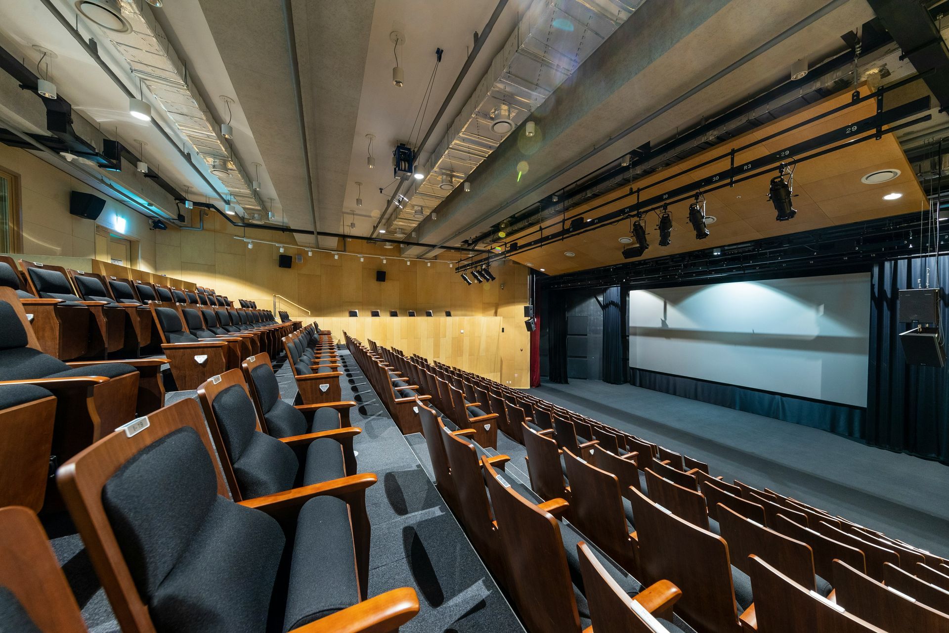 Rows of empty theater seats face a stage with a screen, overhead lights.