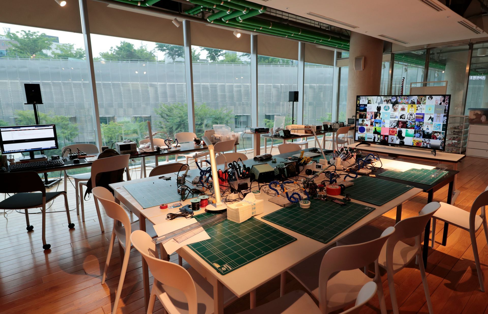 Workshop with tables, tools, and a large screen displaying a video call, near a large window.