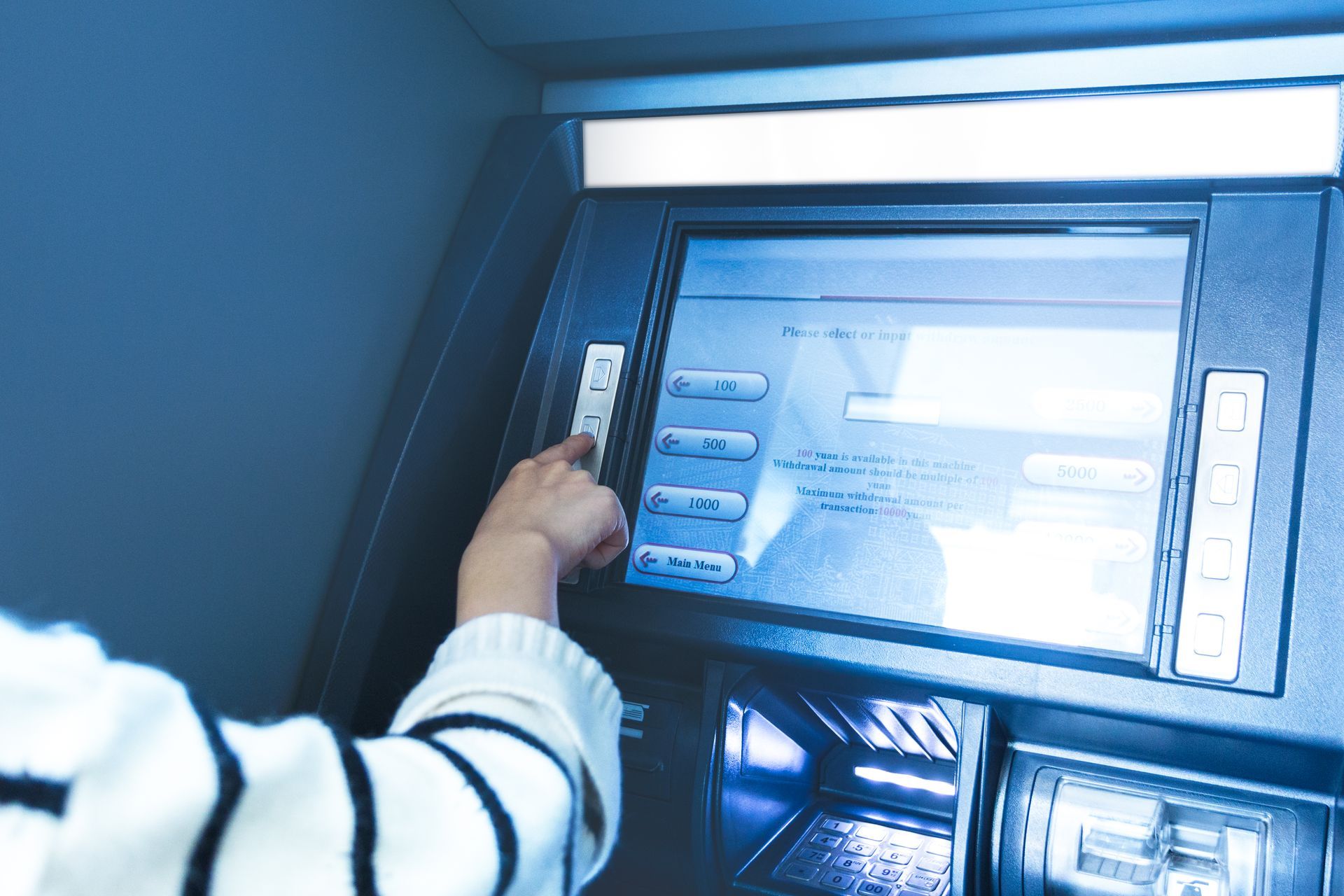 Person using an ATM. Their hand presses a button on the machine's side. Blue-toned image.
