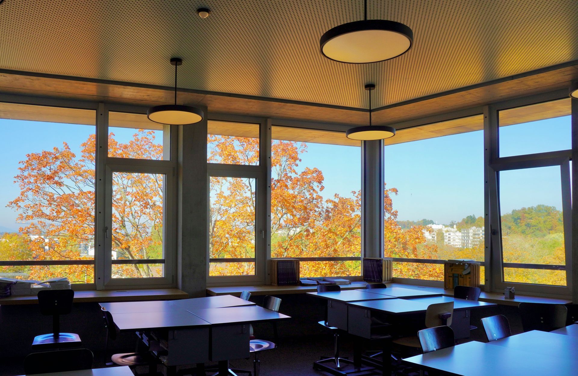 Bright room with large windows overlooking autumn trees, desks, and circular ceiling lights.