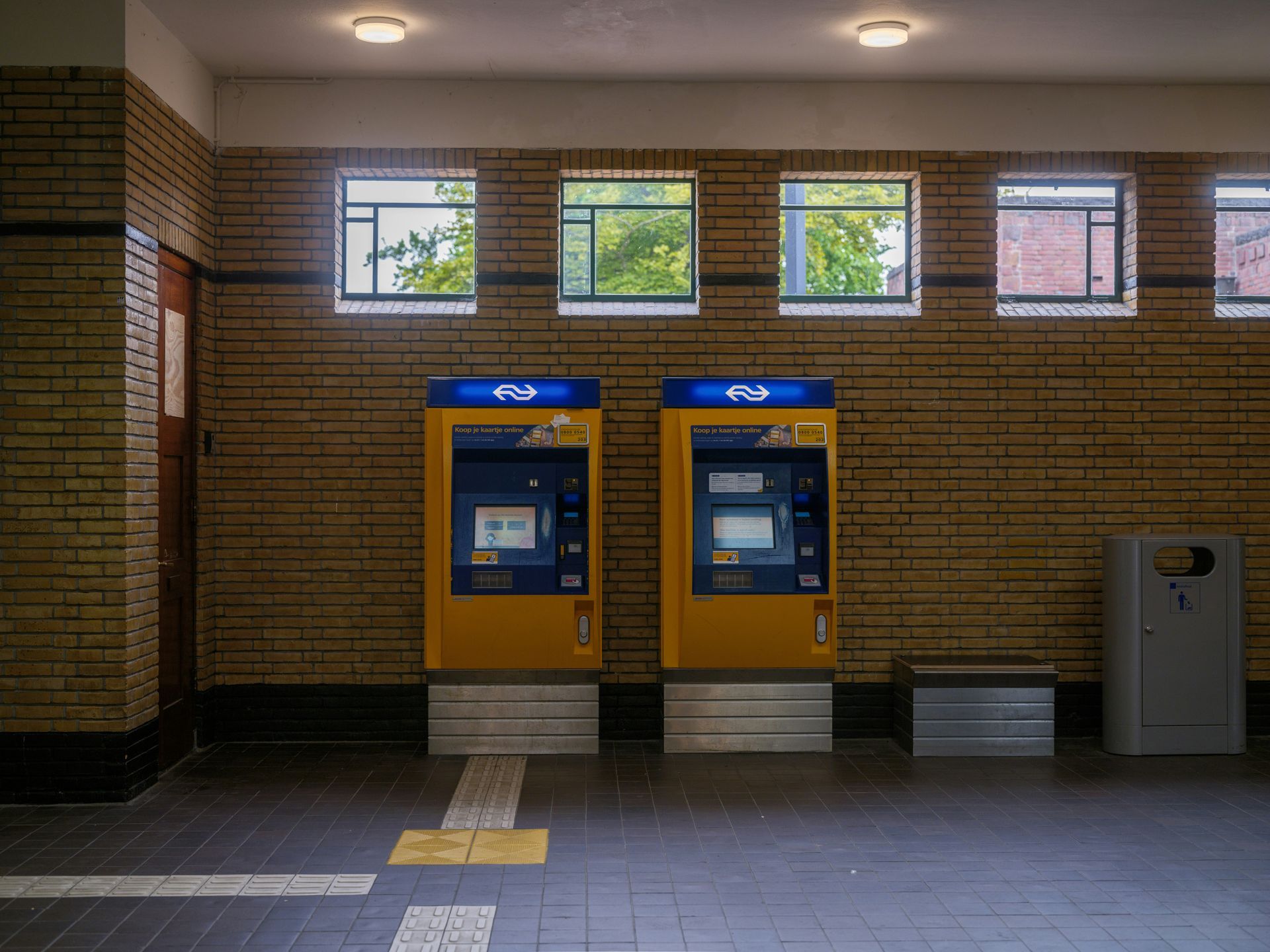 Two yellow ticket machines at a train station against a brick wall, with windows above.