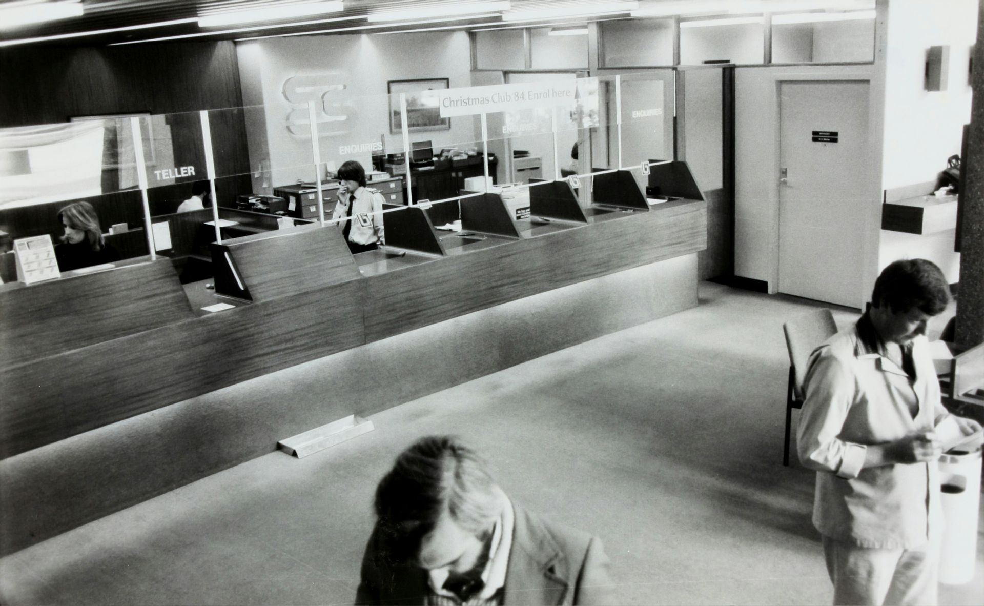 Bank interior with employees behind a counter. Customers stand nearby.