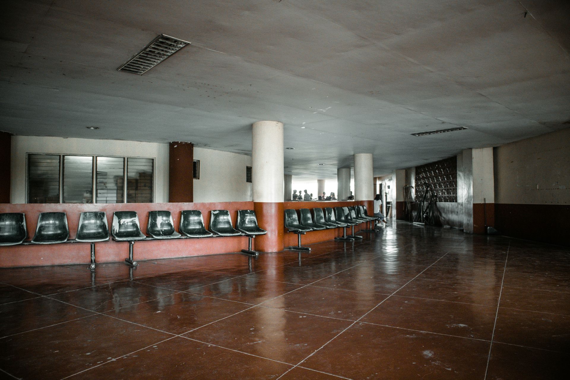 Empty, aging waiting room with rows of chairs, red tile floor, and a white pillar.