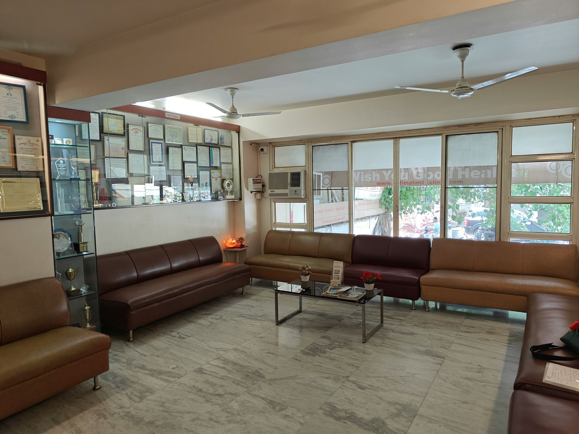 Waiting room with brown leather sofas, glass coffee table, and windows. Wall displays certificates.