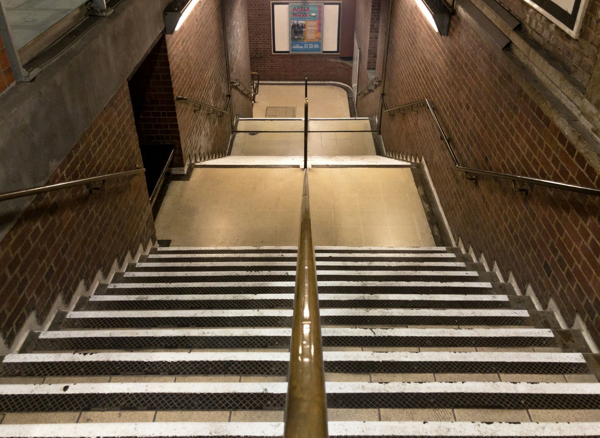 Staircase descending into a subway station. Brick walls and metal handrails flank the steps.