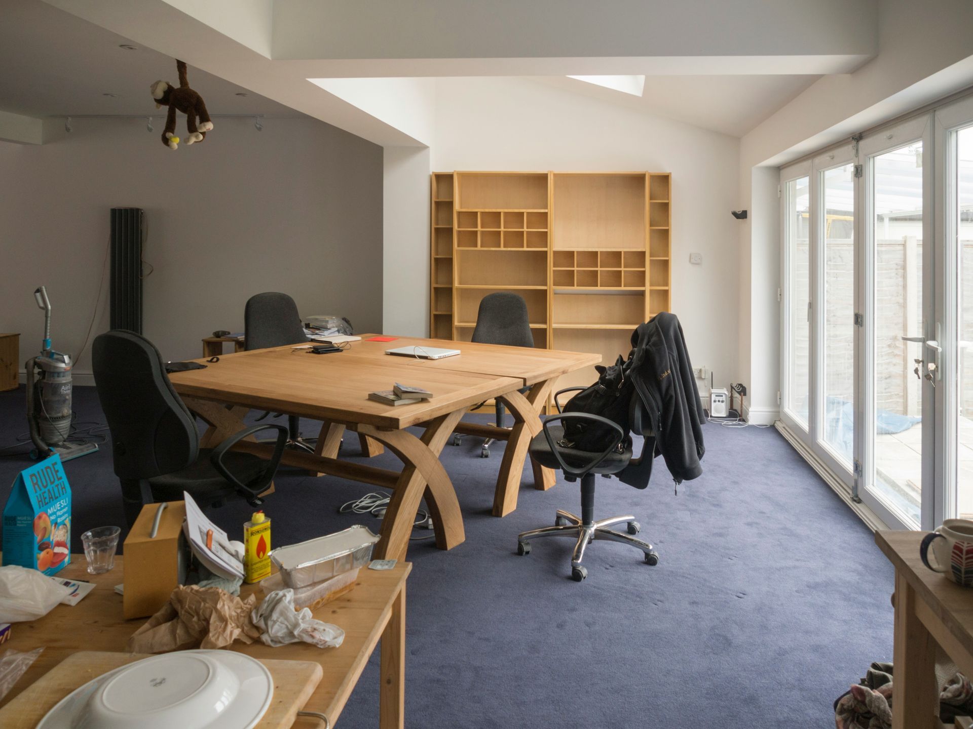 A cluttered home office with a large wooden table, built-in shelving, and blue carpet.