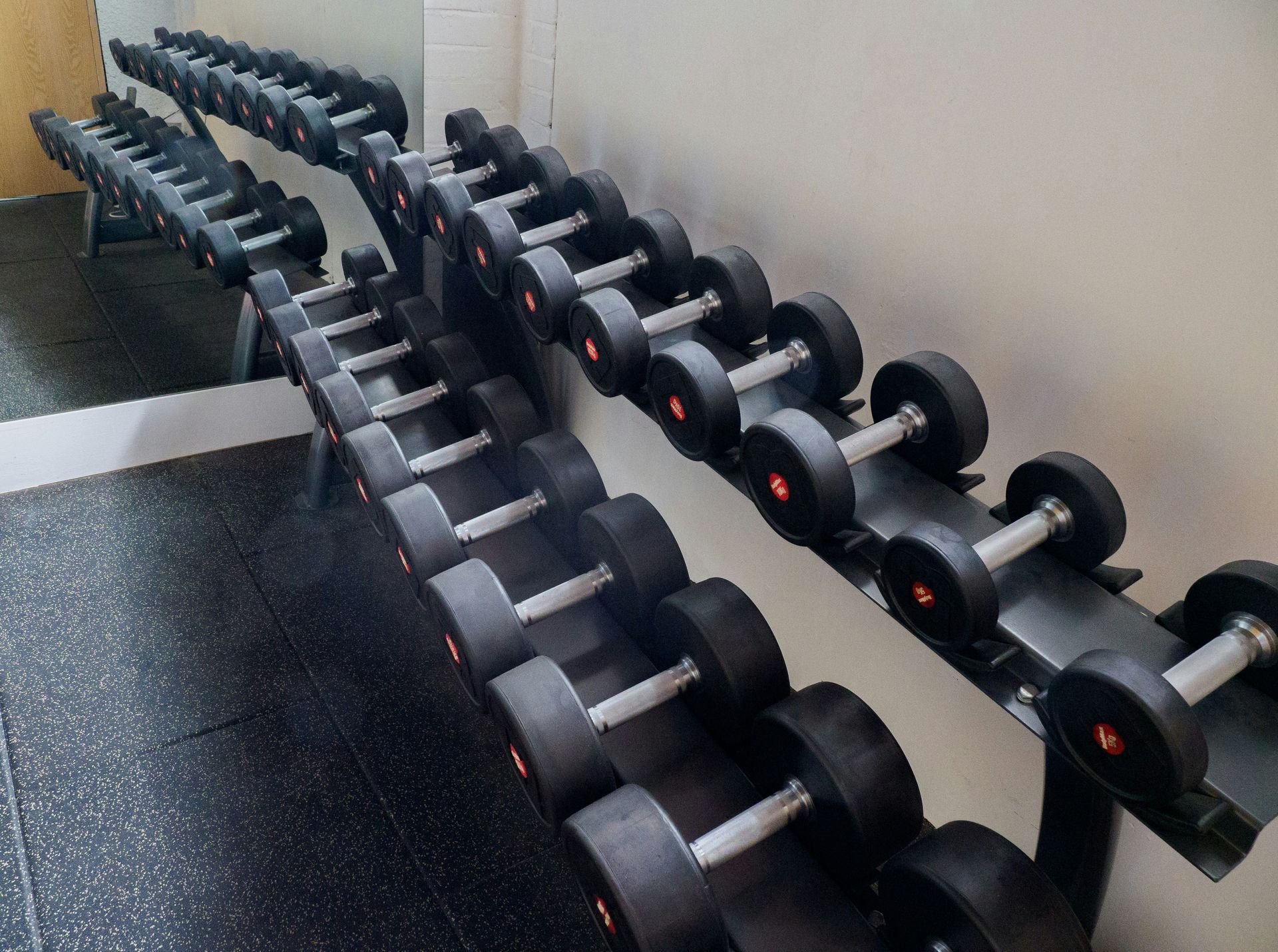 Rows of dumbbells on a rack in a gym, with a mirror reflecting the weights and flooring.