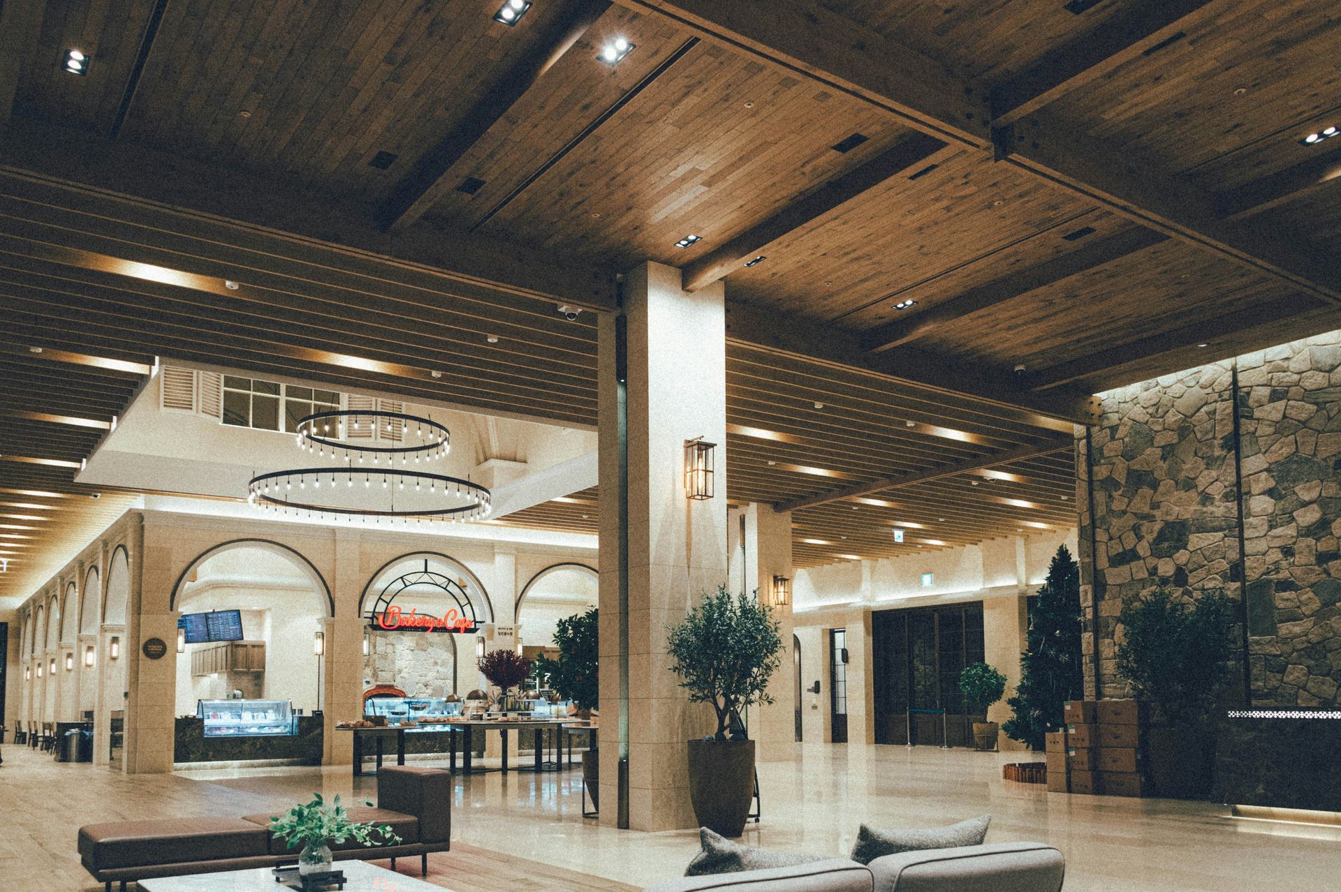 Lobby with wooden ceiling and columns, stone wall, and plants. Hallway with archways in the background.