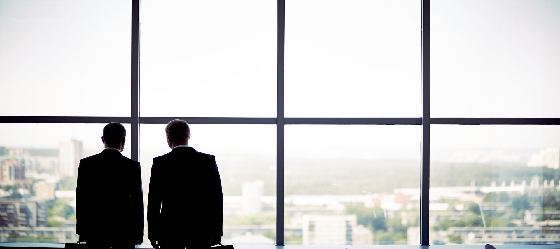 Two businessmen in suits look out a large window at a city view.