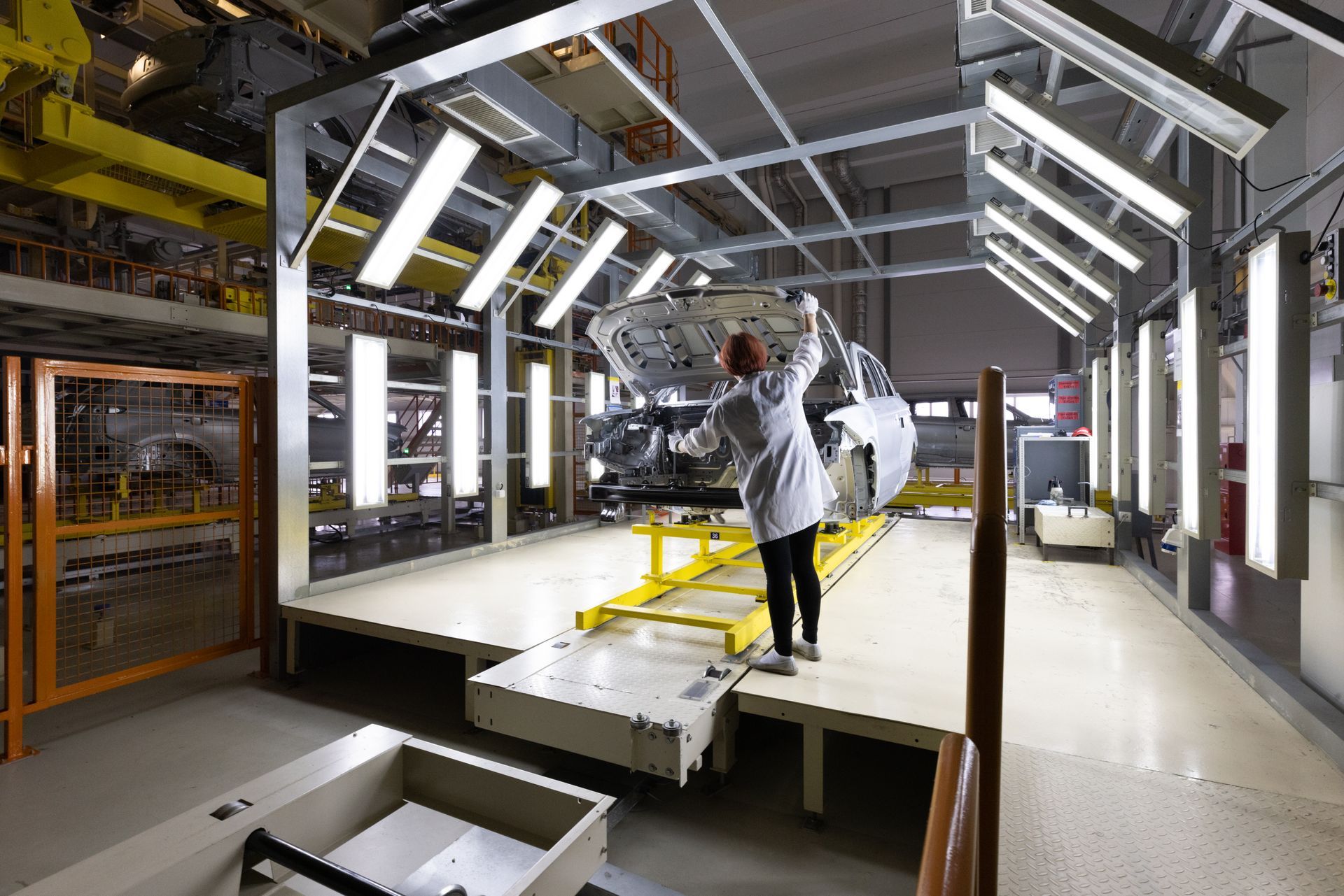 A person in a lab coat inspecting a car body in a brightly lit factory.