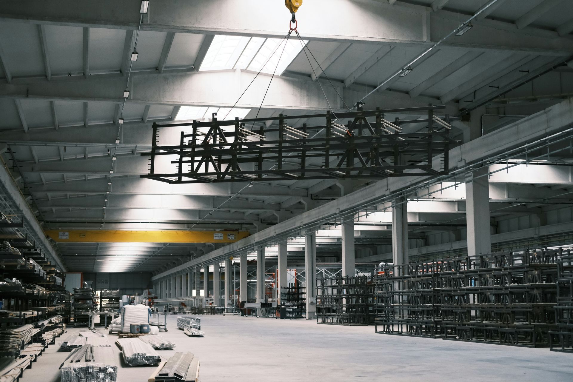 Factory interior with a metal structure suspended by an overhead crane, sunlight through skylights.
