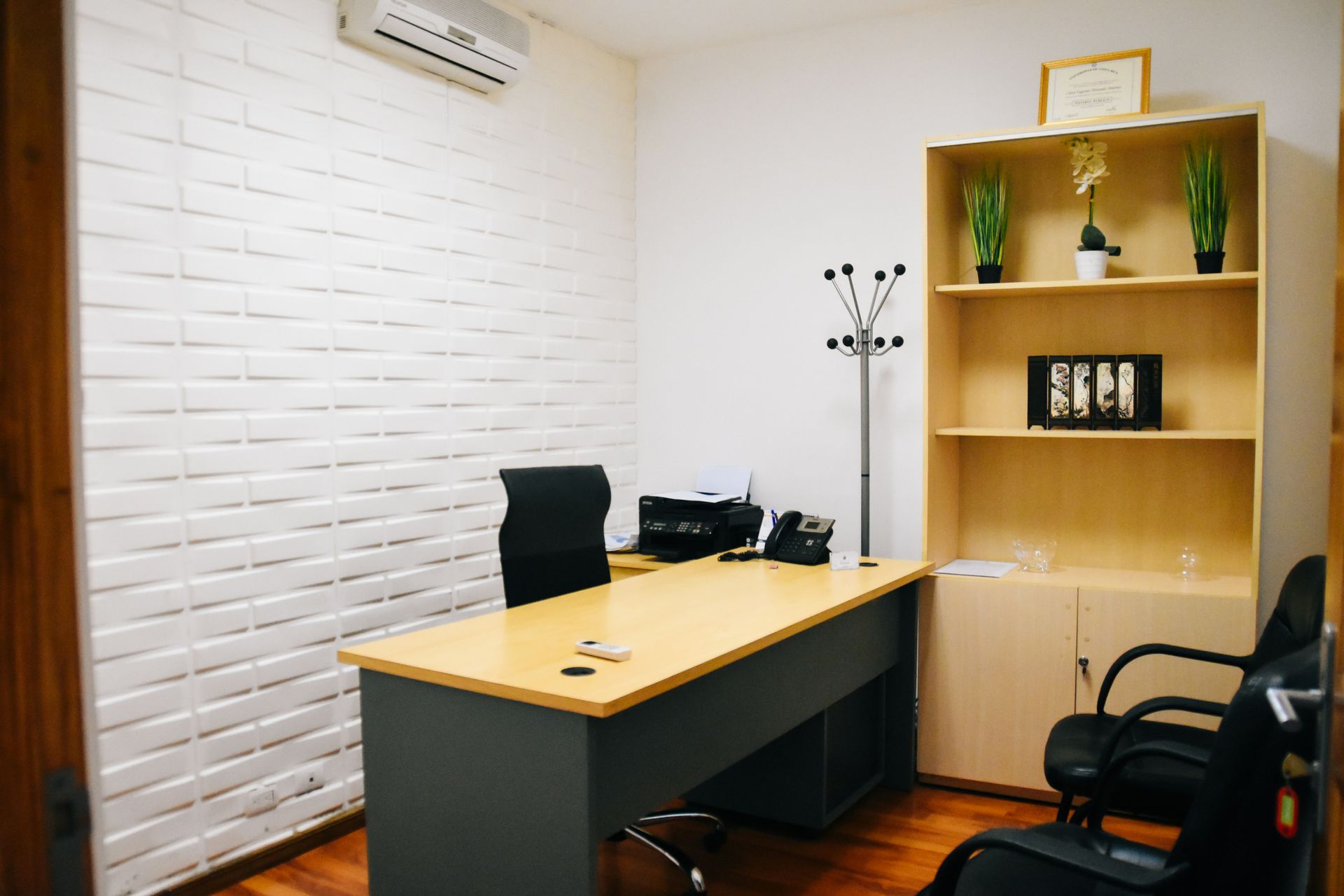 Office with desk, shelving unit, brick wall, chair, and coat rack; light wood and neutral tones.