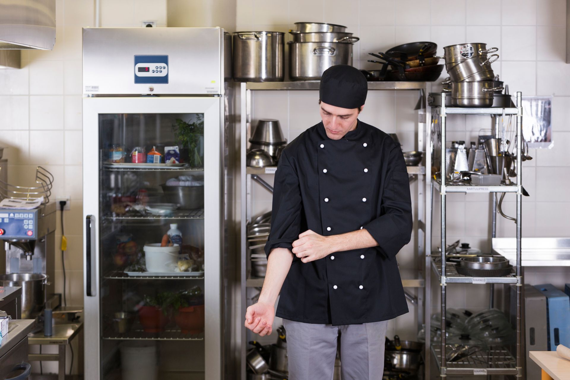 Chef in black uniform rolling up sleeves in a commercial kitchen with stainless steel appliances.