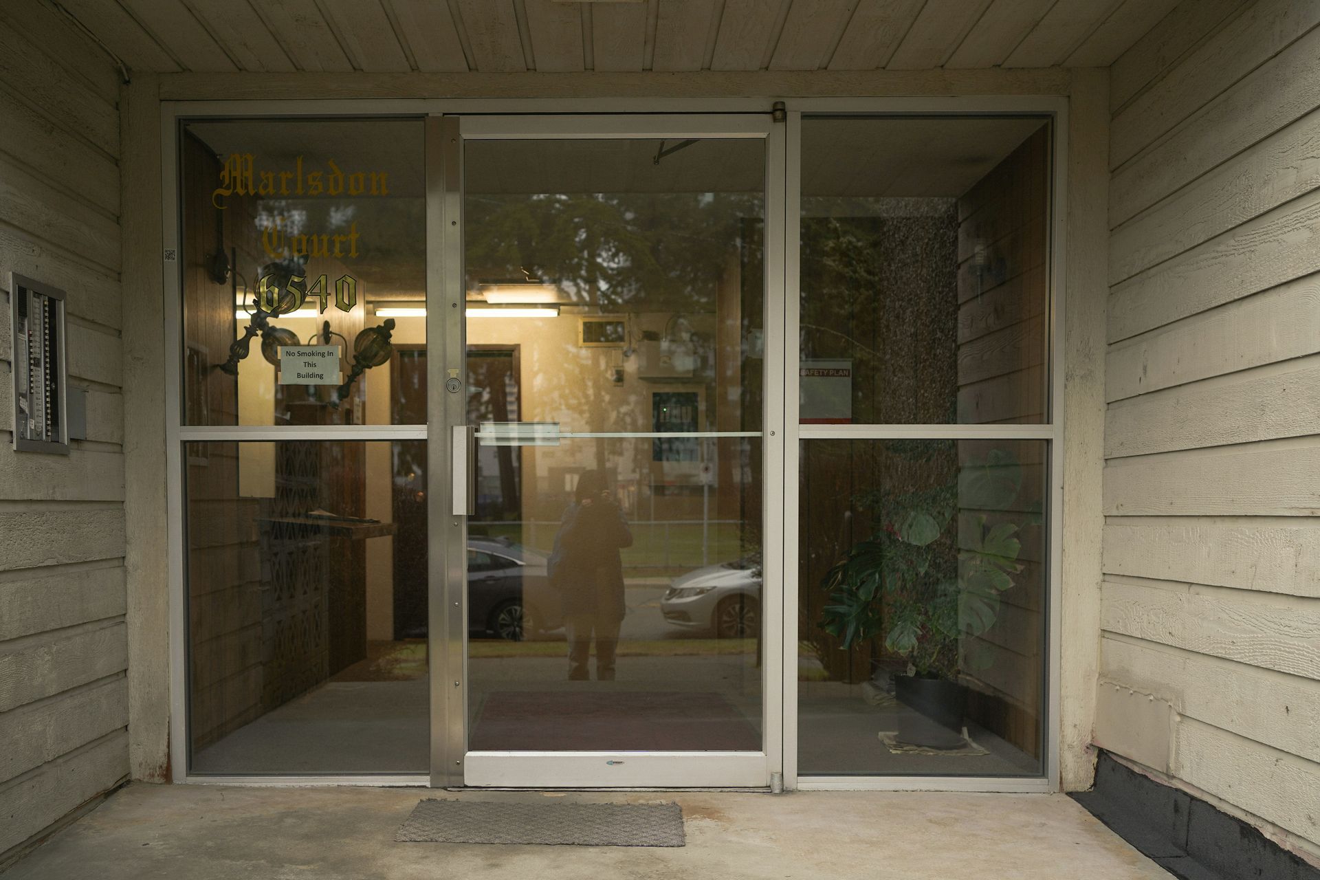 Exterior view of a building entrance with glass doors, a person visible inside, and off-white siding.