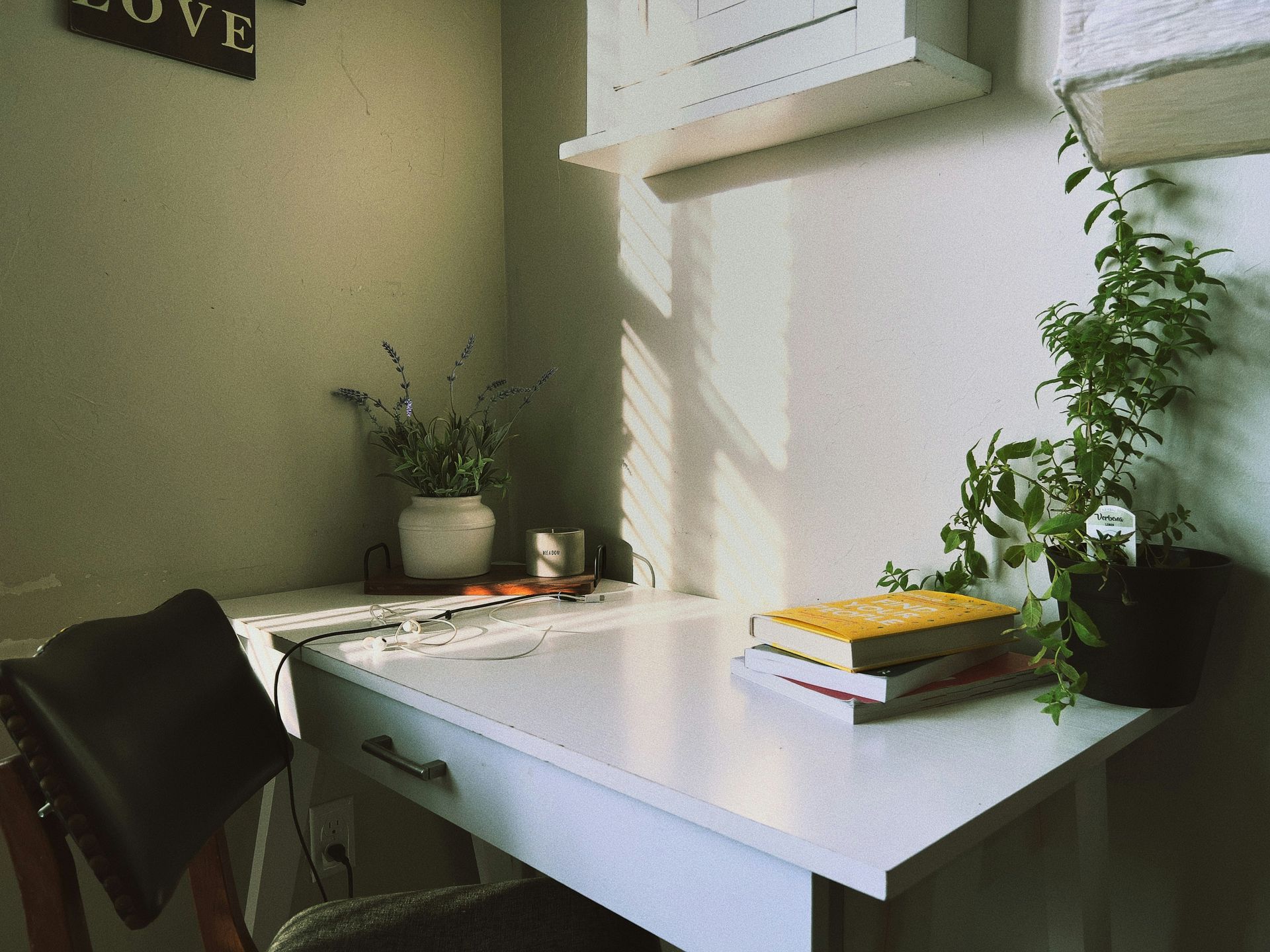 White desk in a corner, with books and a plant. Sunlight streams through blinds.