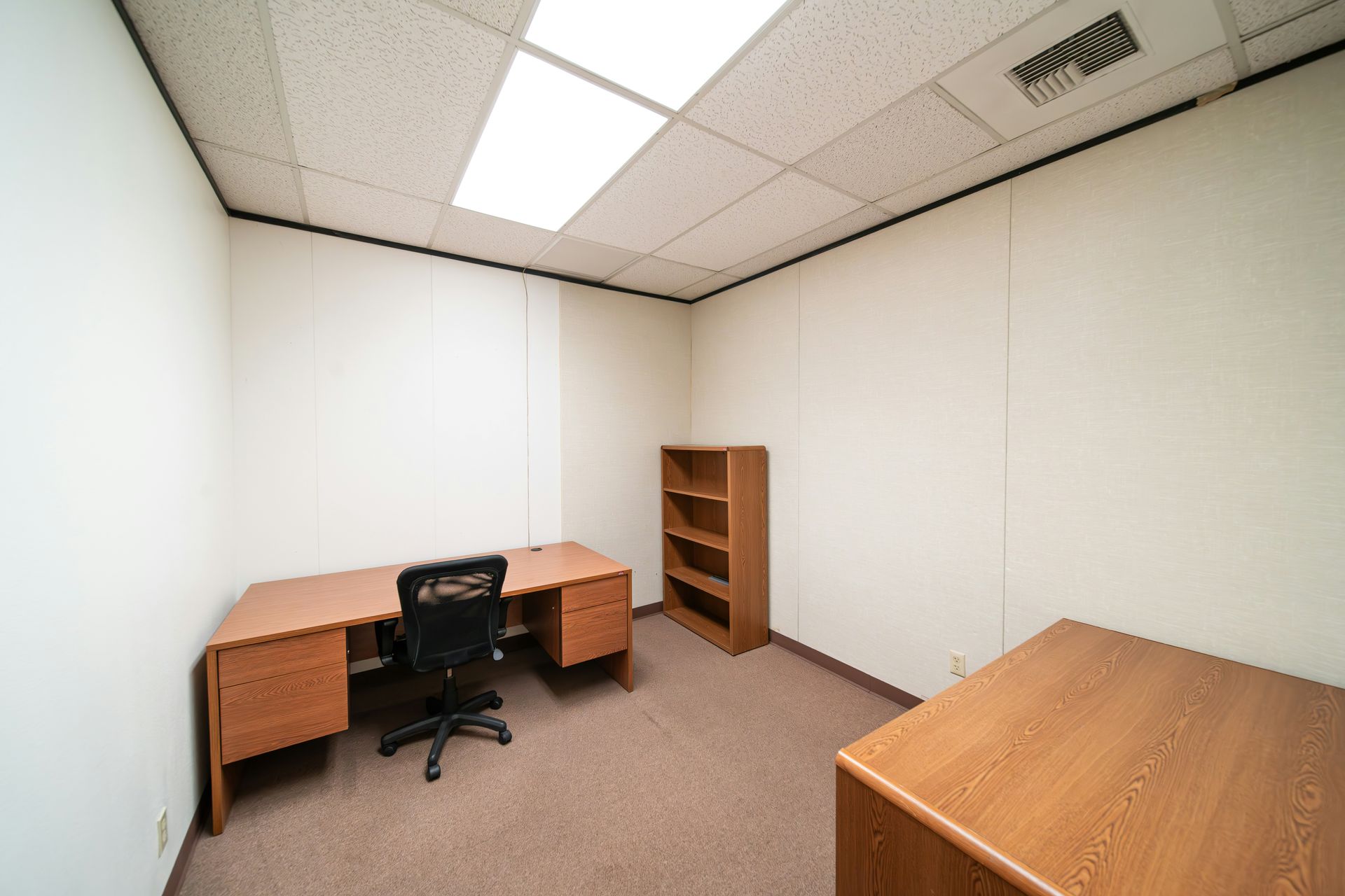 Empty office with desk, chair, bookcase, and a second table. Neutral walls, brown carpet.