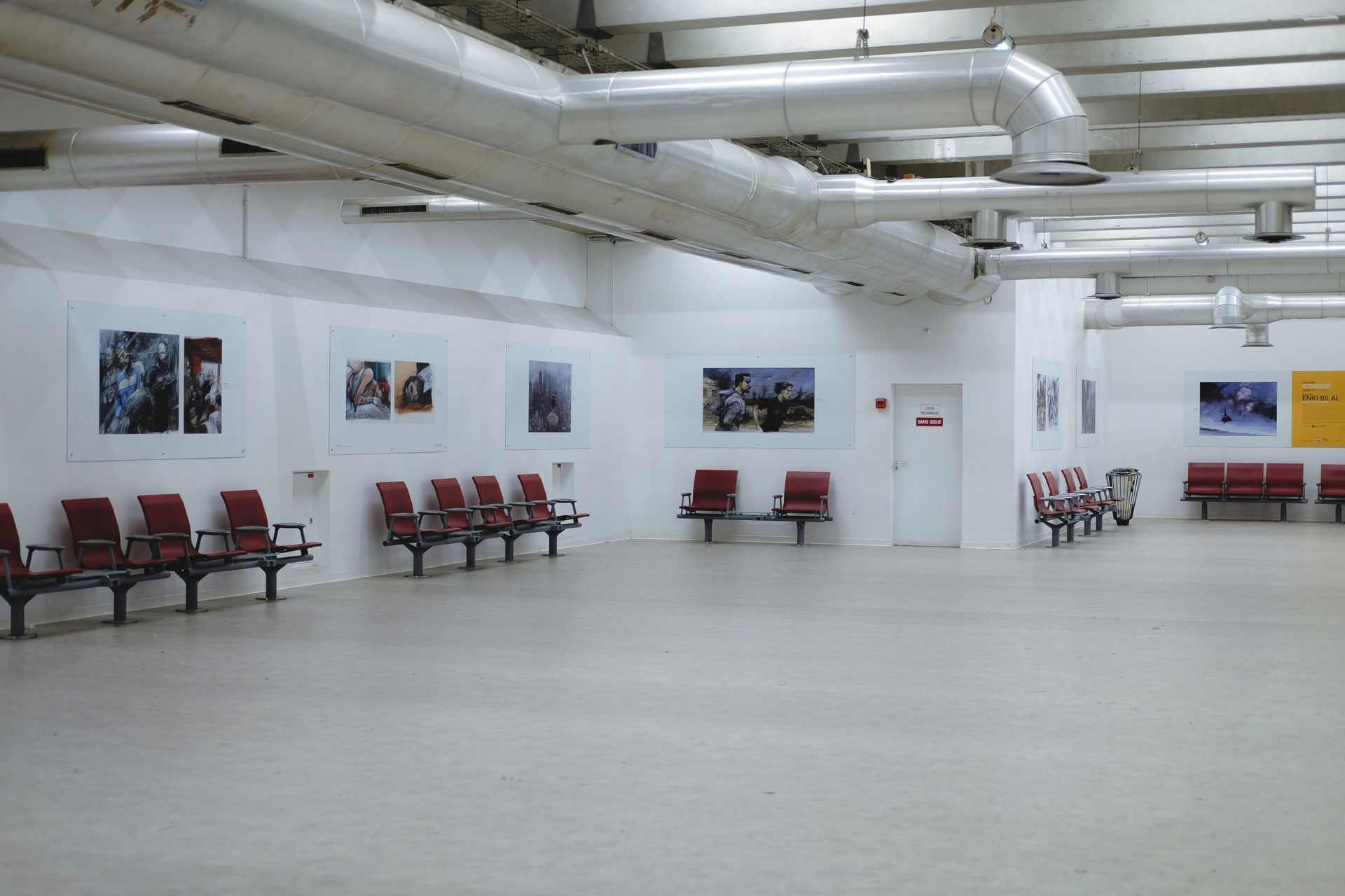 Empty gallery space with red seating, white walls, and artwork. Overhead ventilation ducts.