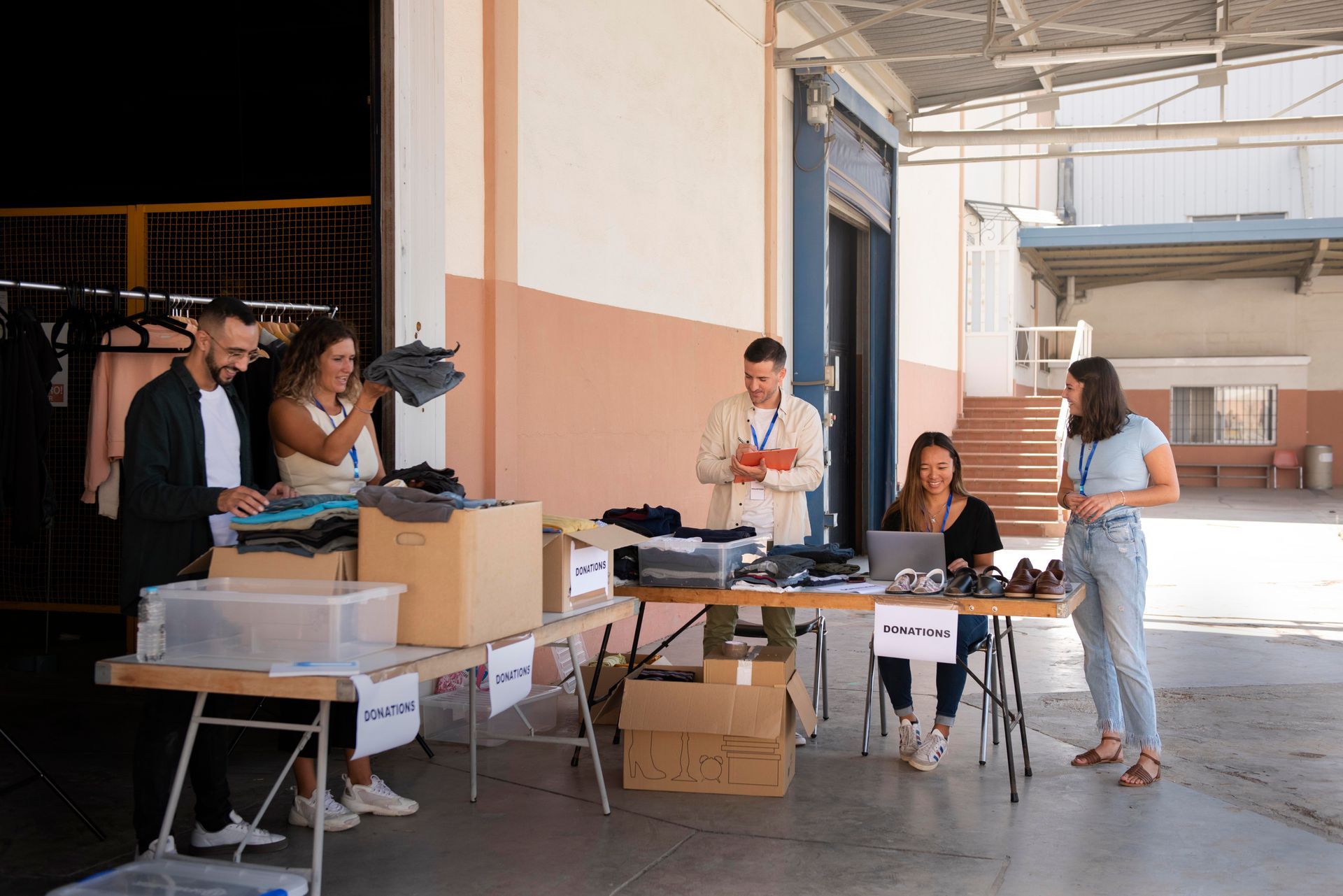 People organize clothes at a table outdoors. Cardboard boxes, racks of clothing, and an open doorway are in view.