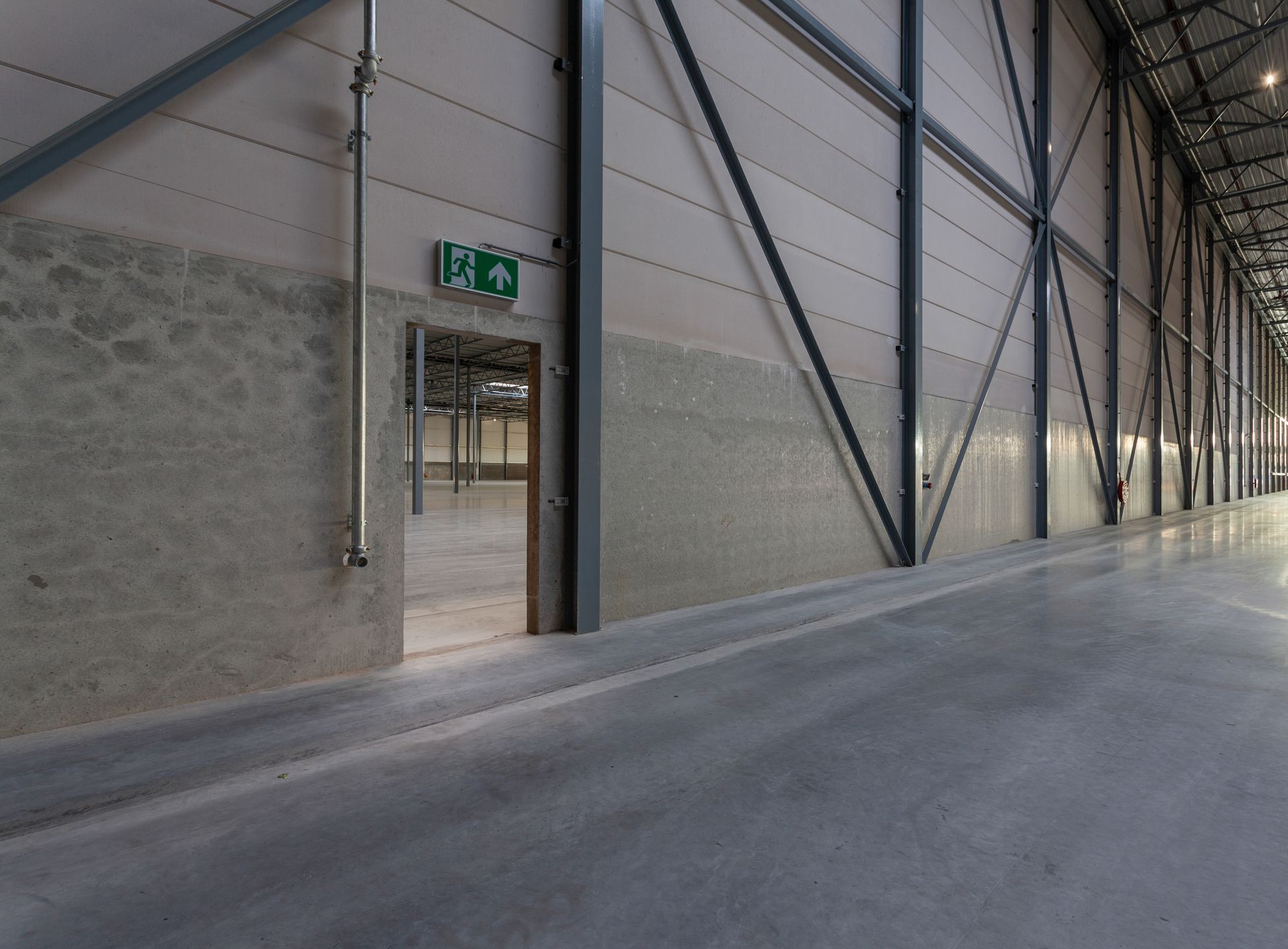 Interior of a large industrial building, concrete floors, metal supports, exit door with green emergency sign.