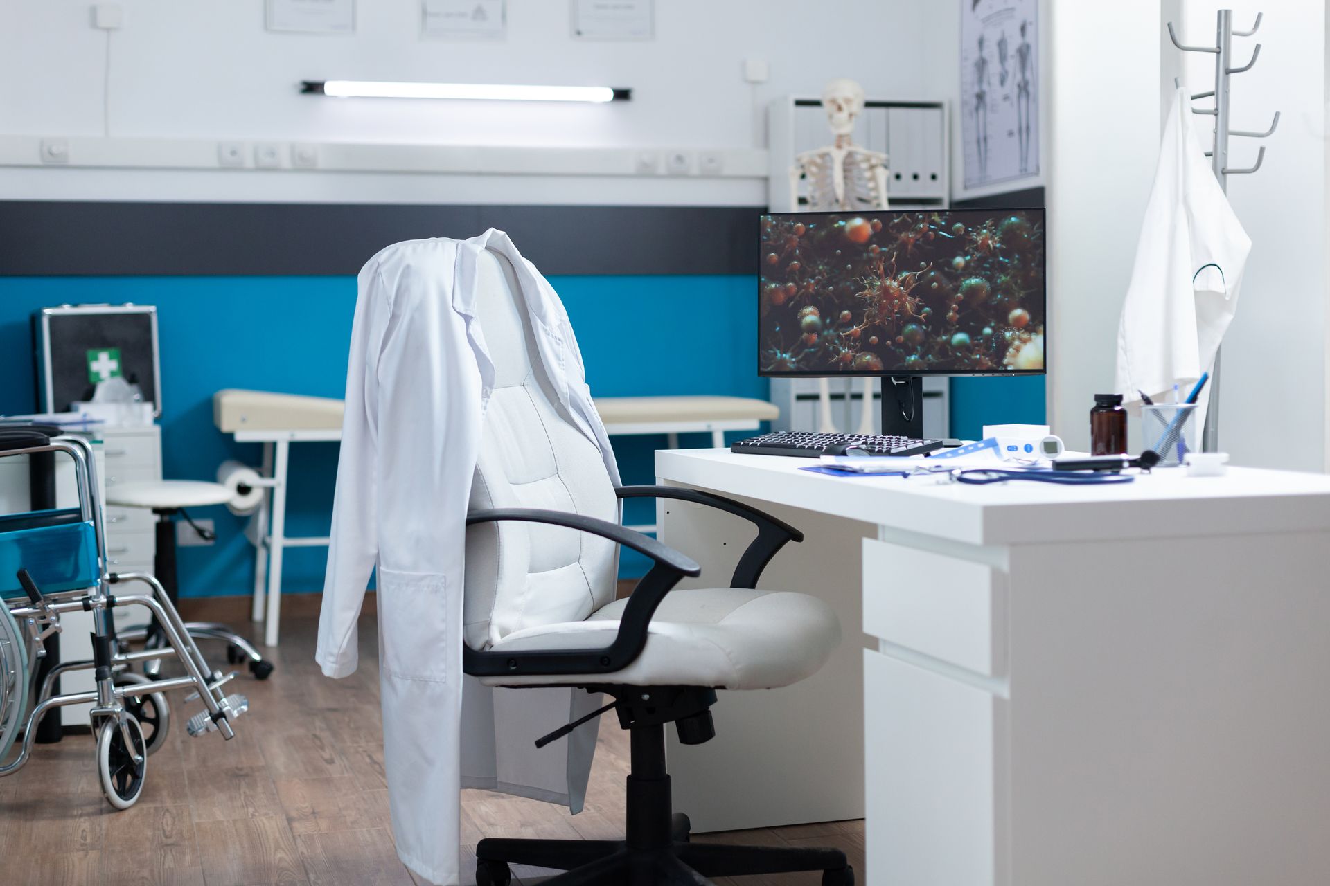 Doctor's office interior with desk, chair, lab coat, wheelchair, and medical equipment.