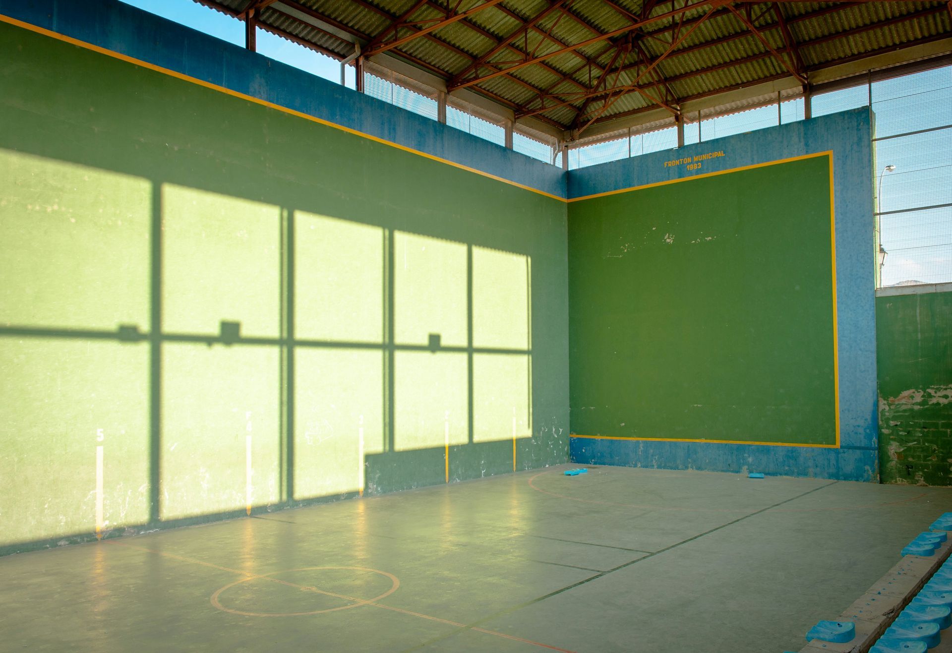 Empty indoor handball court with green and blue walls, glass windows, and concrete floor.